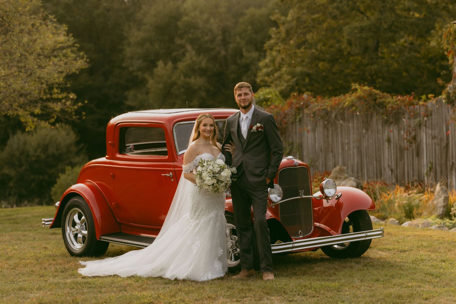 A romantic wedding photo session featuring a vintage red car in a rustic outdoor setting during golden hour.