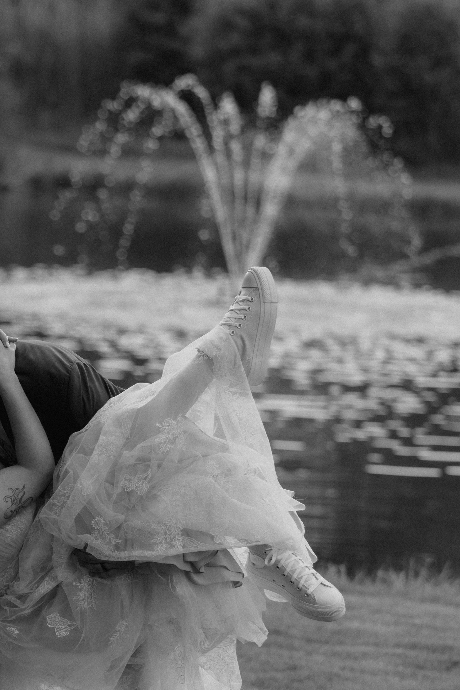 A person lounging by a lake lifts their leg adorned in ballet shoes and tulle, with a water fountain spraying in the background.