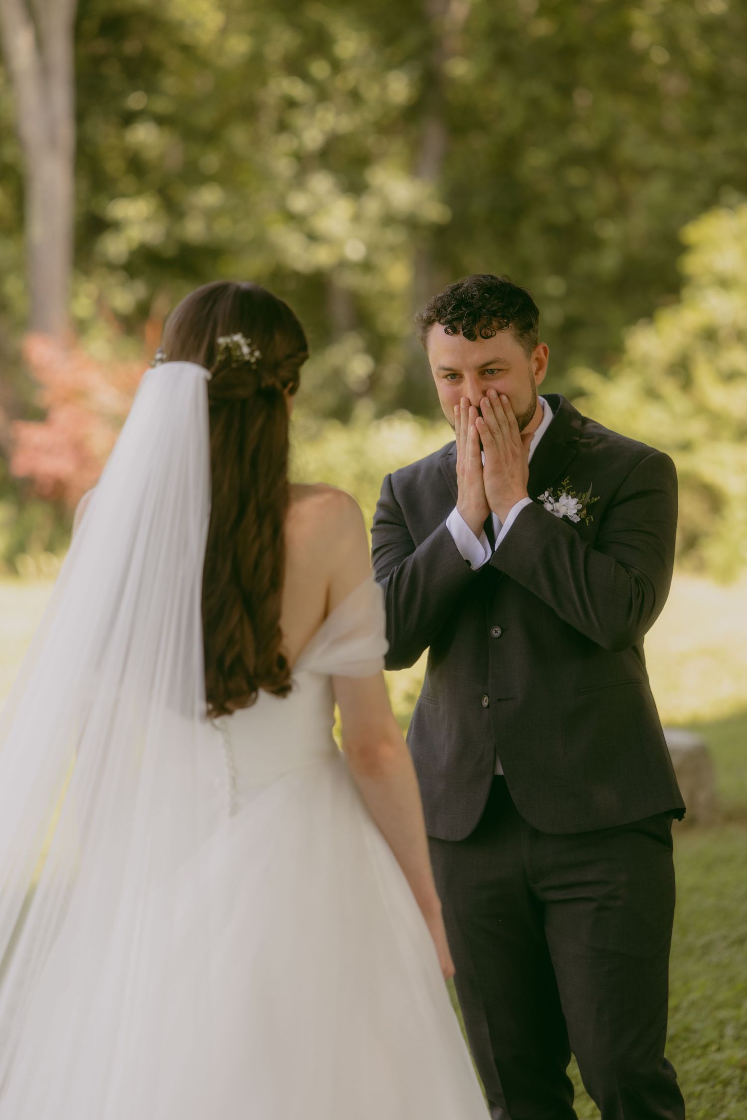 Emotional first look moment captured during outdoor wedding ceremony as groom covers face while seeing bride in gown and veil.