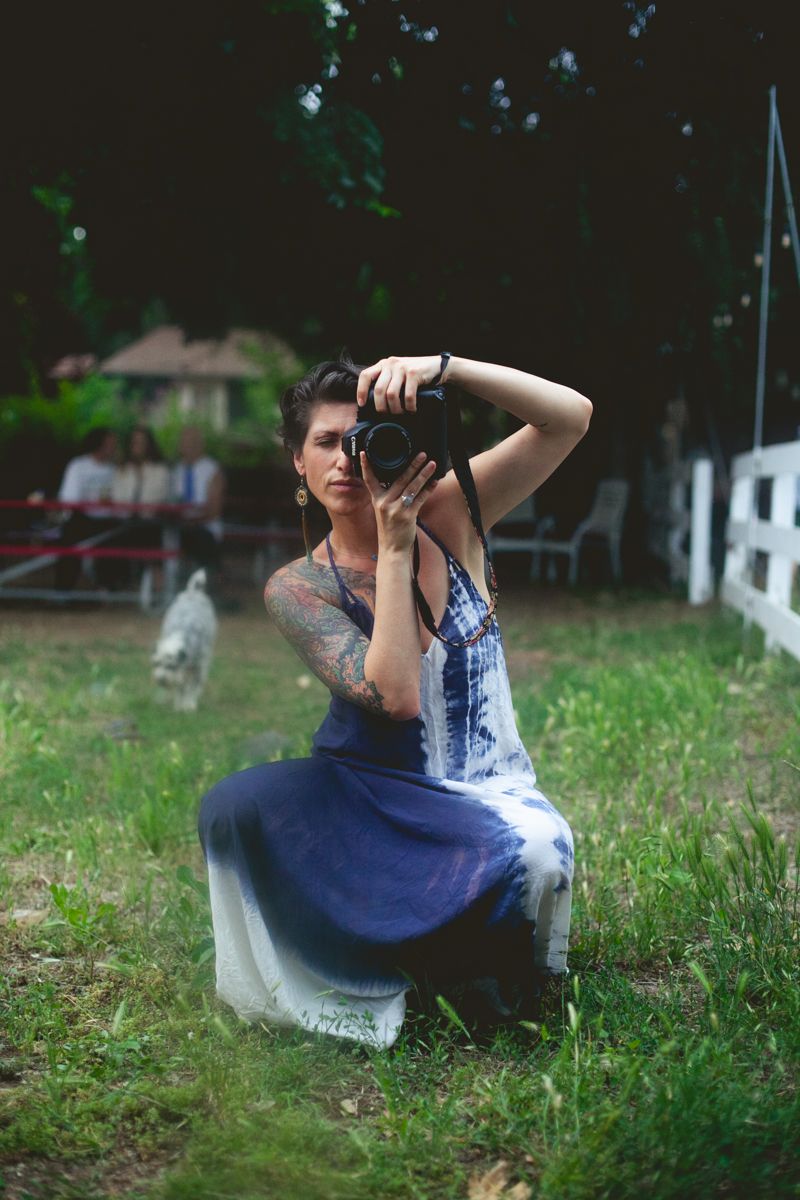 Photographer in blue dress crouches in grass while taking photos near a white fence.