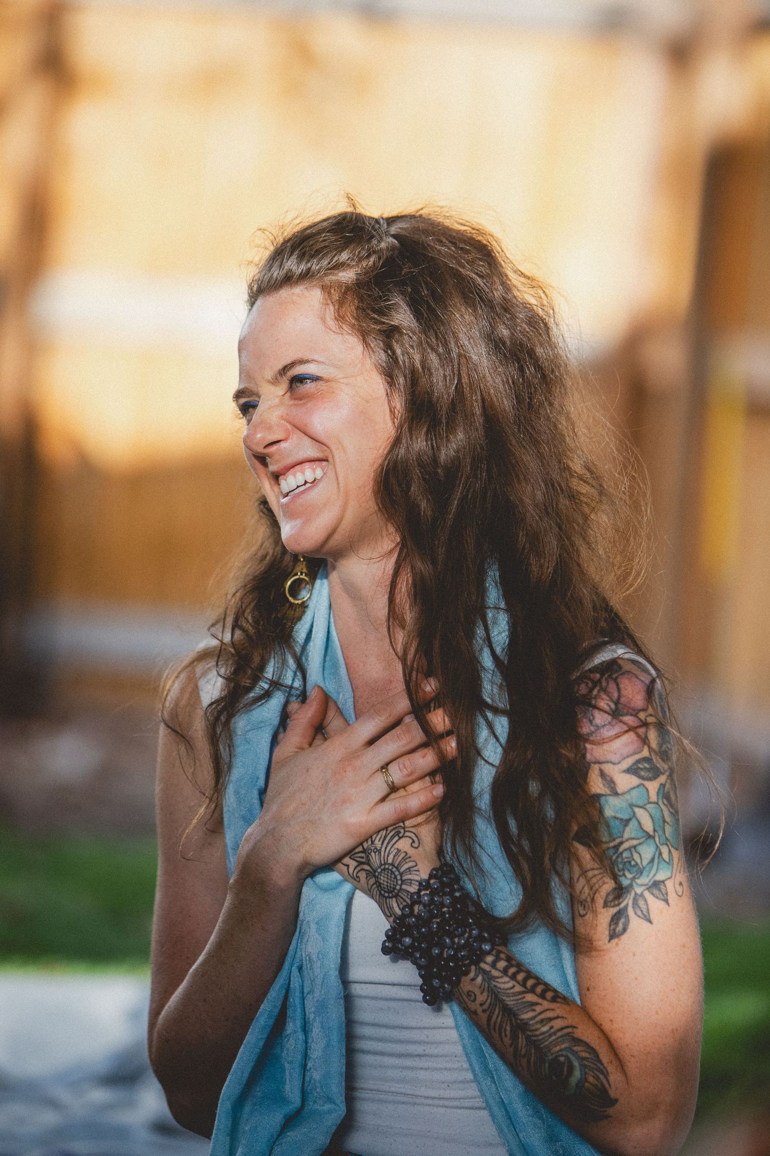 Woman with long dark curly hair and blue blouse laughing joyfully in golden sunlight outdoors.