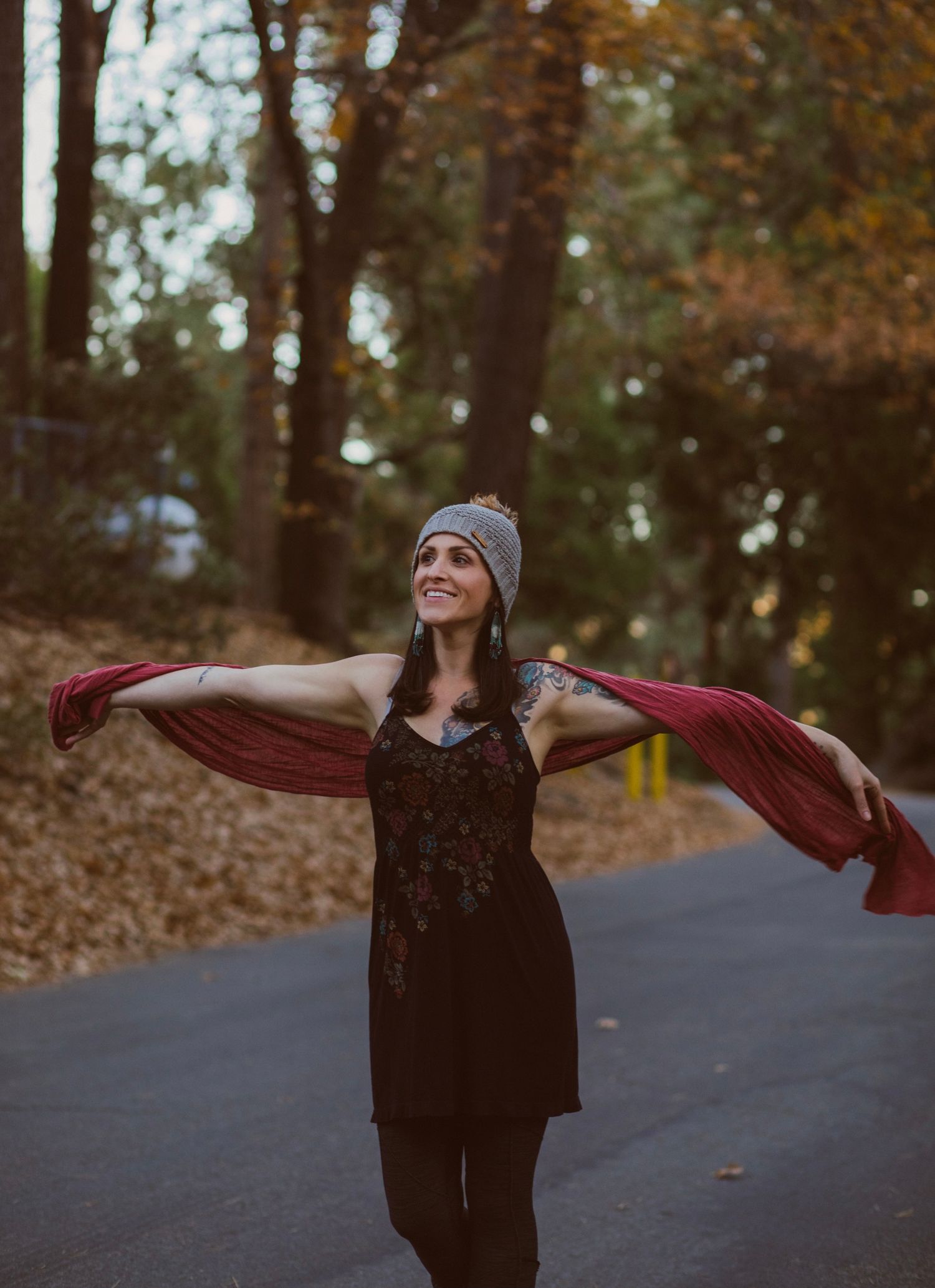 Person in black dress and beanie spreads red scarf while dancing on autumn forest path.