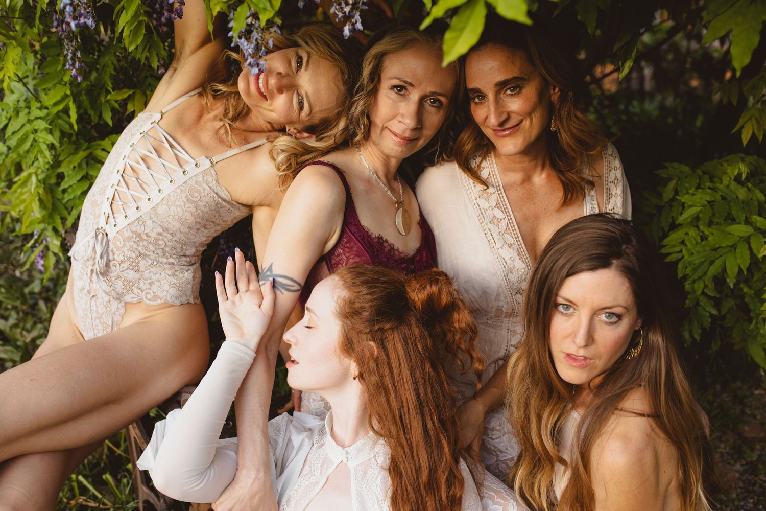 A group of women in light dresses pose together outdoors against greenery.