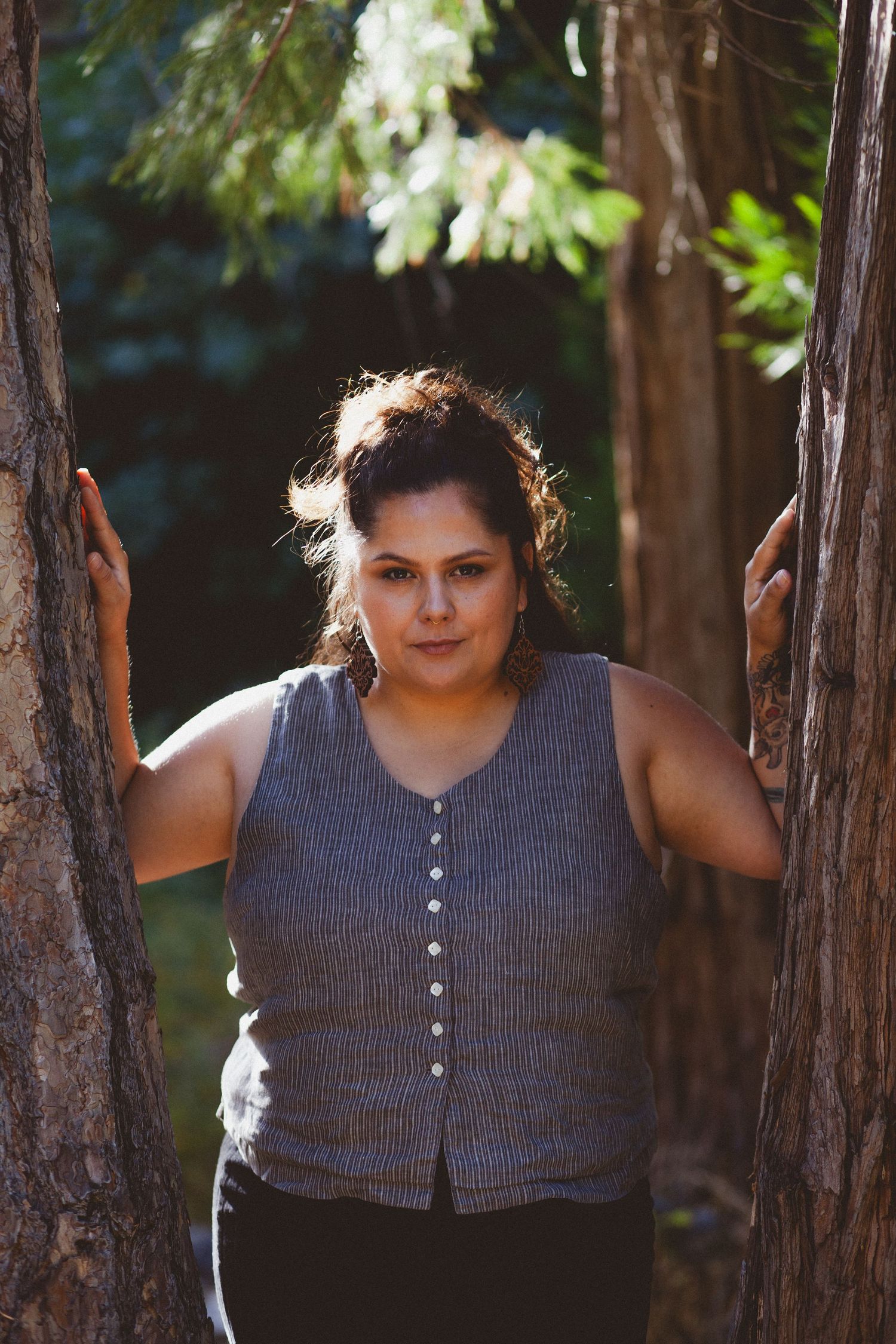 Casual portrait between tree trunks wearing grey button-up vest in forest setting.