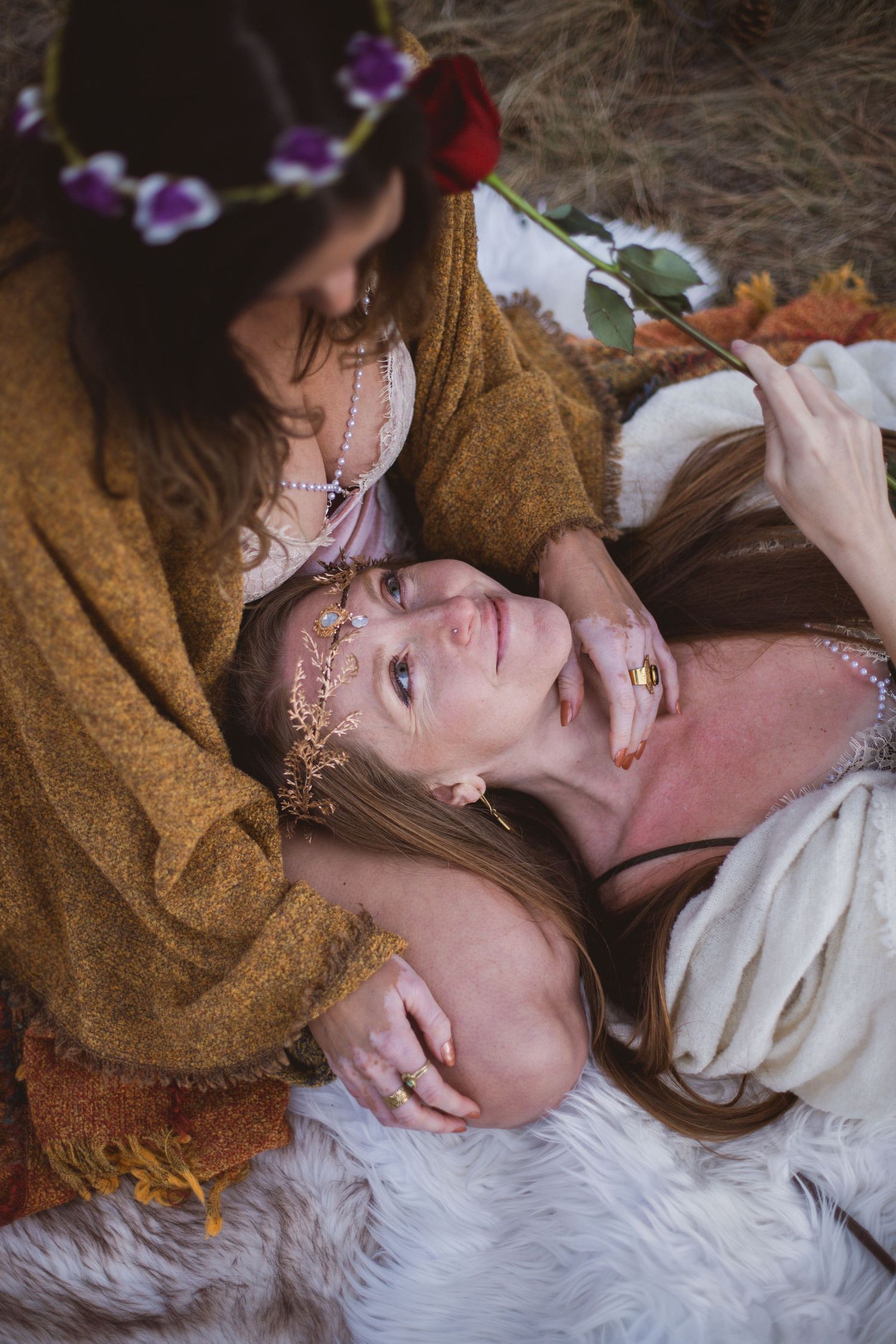 Two people in medieval fantasy inspired costumes lying on white fur in a dreamy ethereal setting with purple flowers.