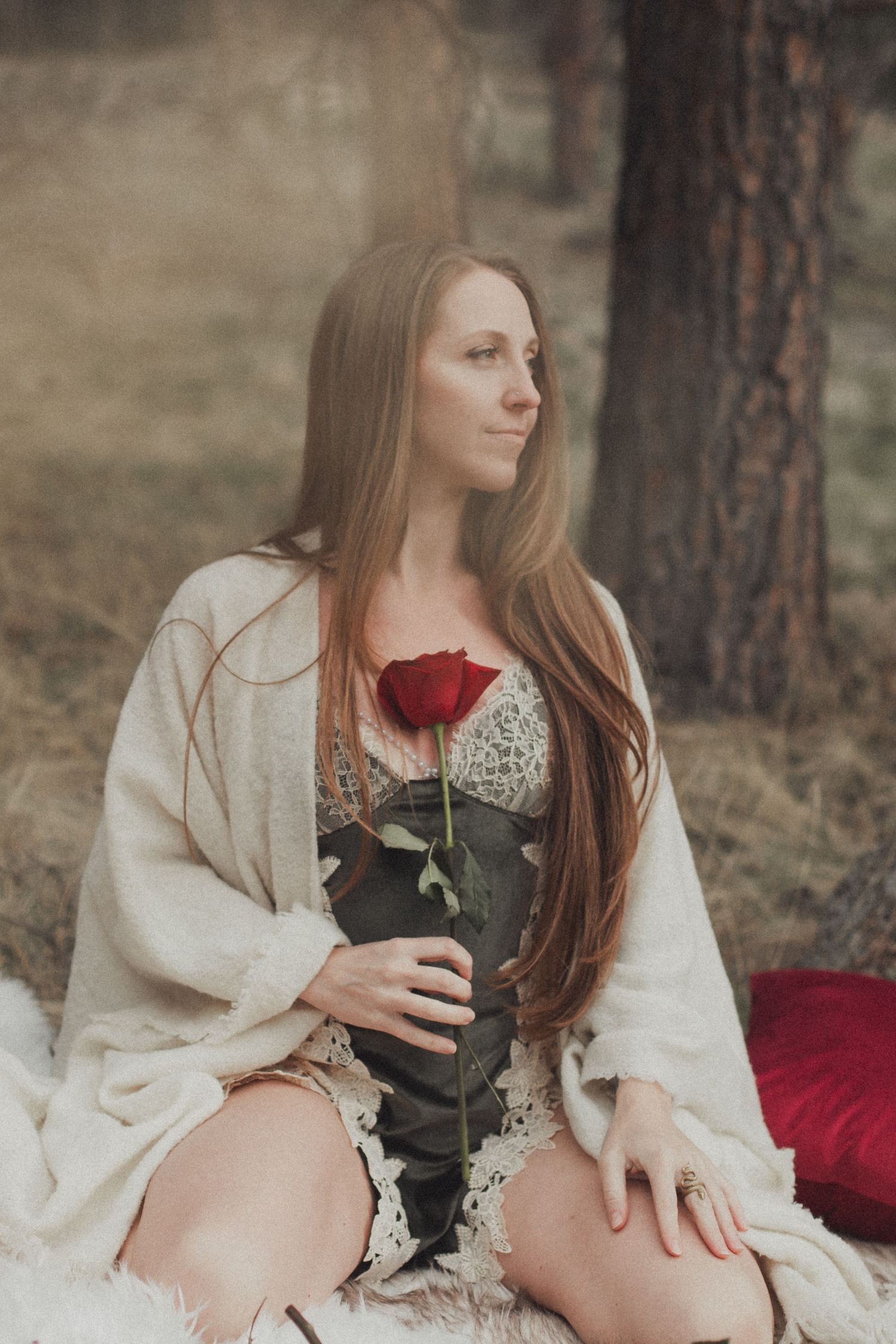 A person in a white cardigan holds a single red rose while sitting on a blanket in a forest setting.
