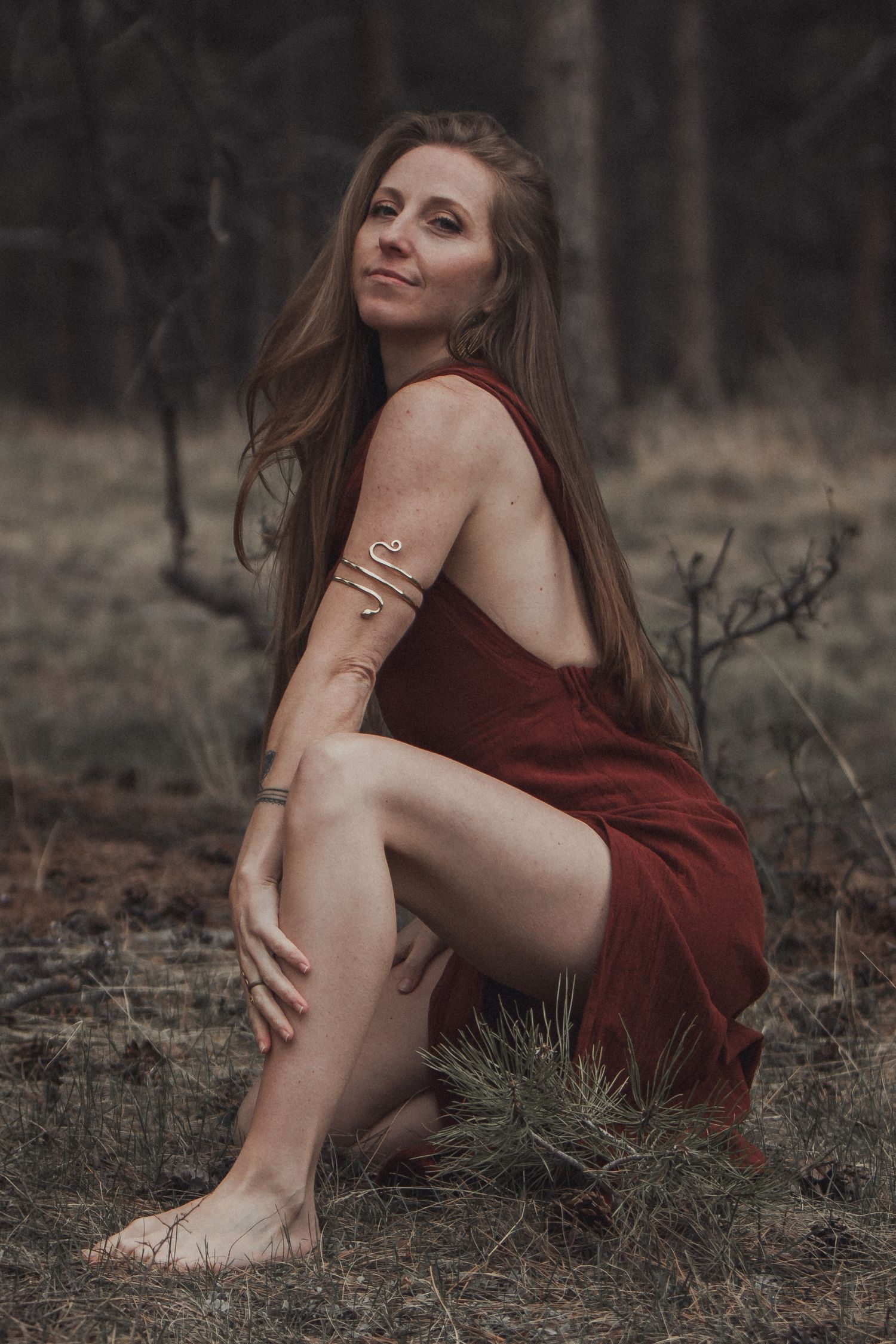 Artistic outdoor portrait of someone in a burgundy dress sitting on autumn ground with metallic arm jewelry.