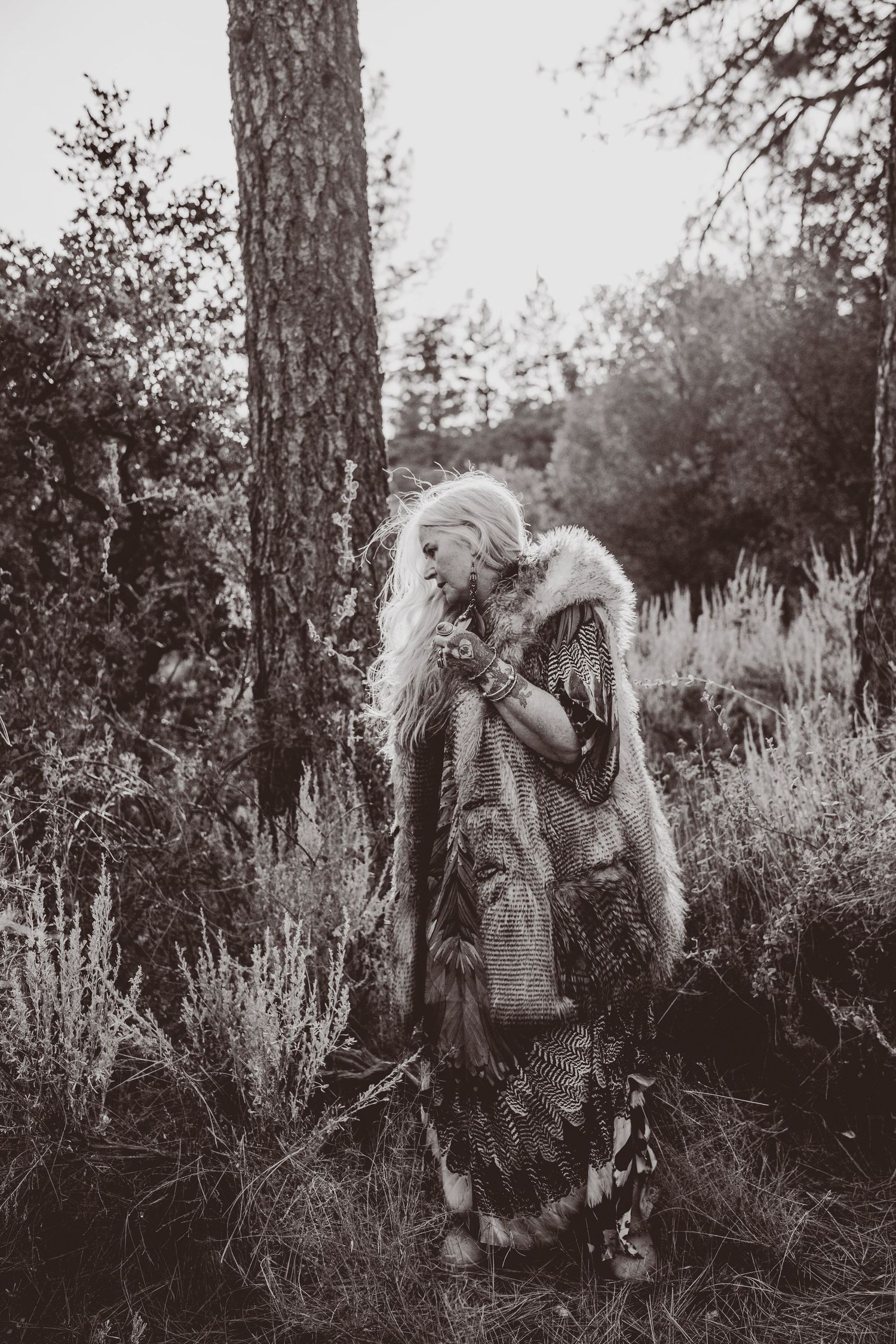 Ethereal figure in fur coat standing among trees in moody black and white forest setting.