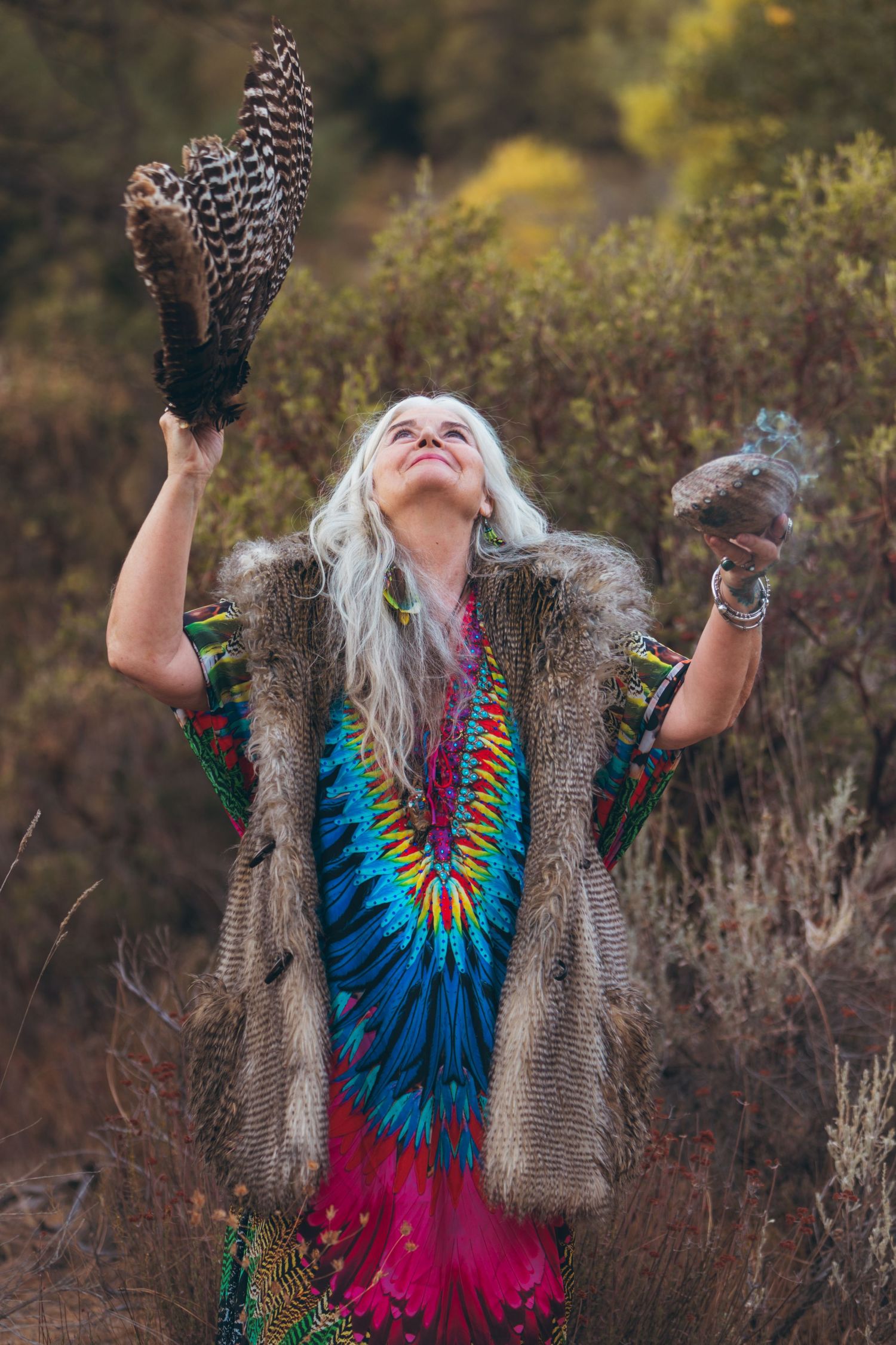 Person in tie-dye dress and fur vest holds feathers up to sky in spiritual gesture.