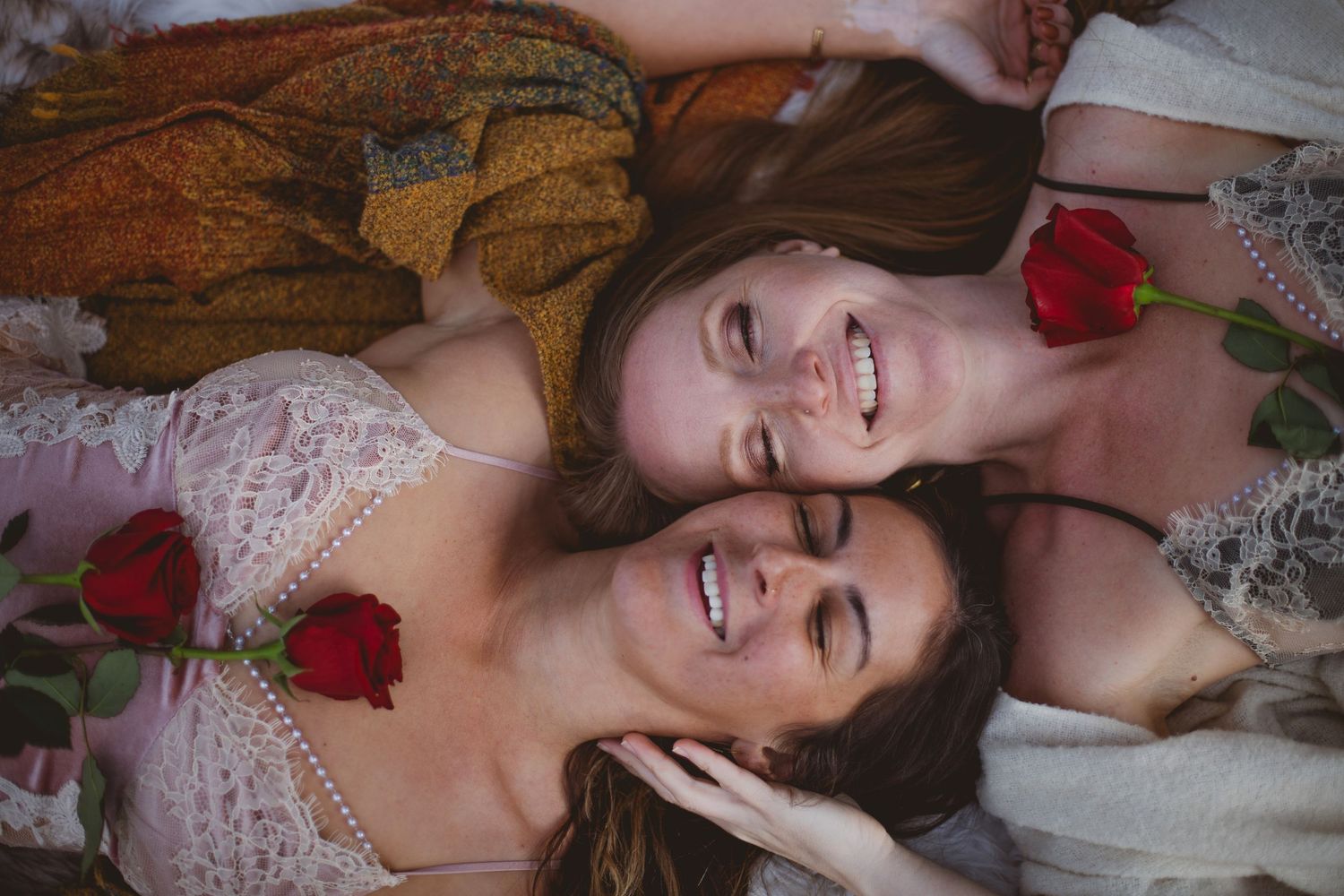 Two people in white lace outfits lying down with red roses and laughing joyfully together.