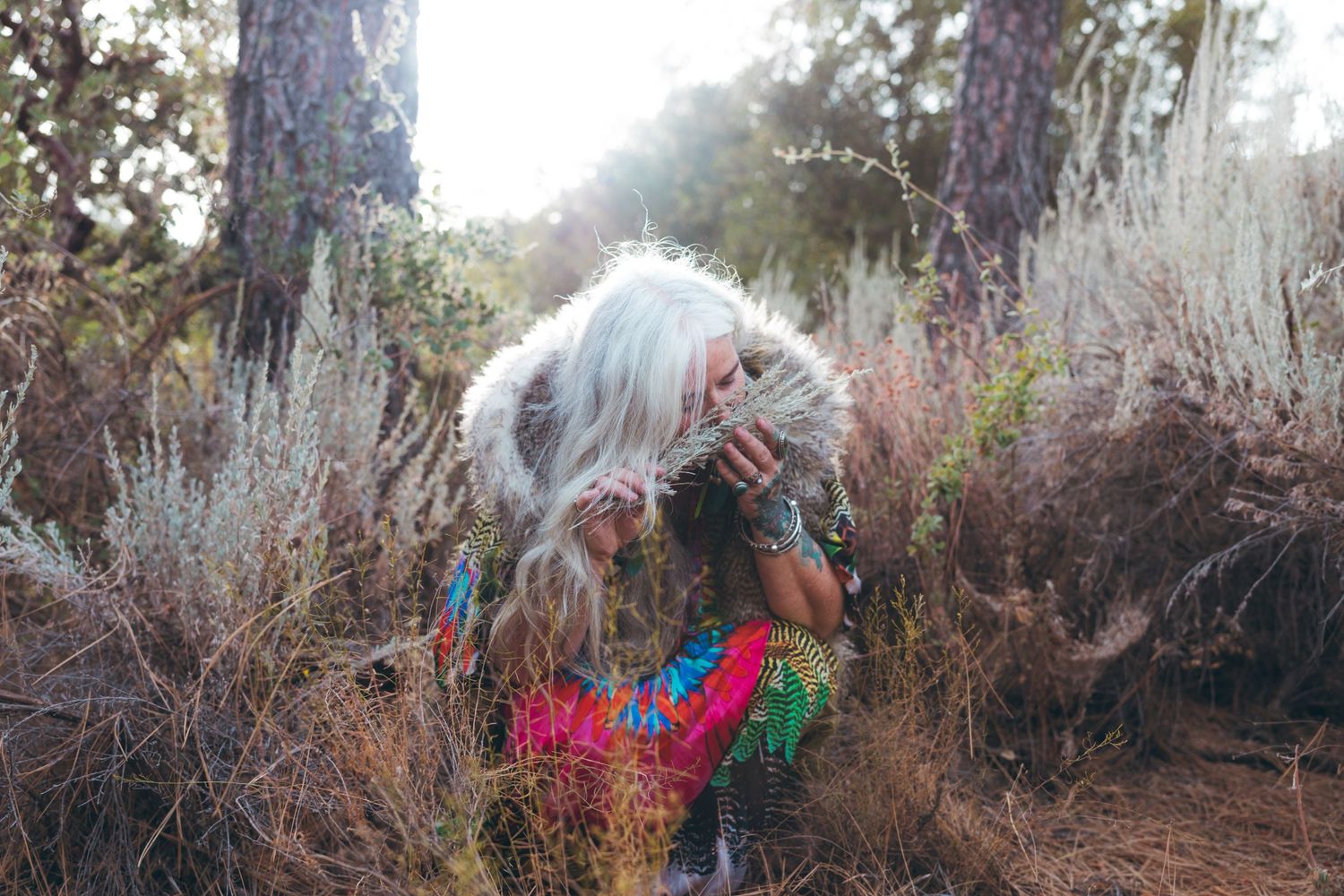 Person with flowing hair stands among dry brush in sunlit forest setting.