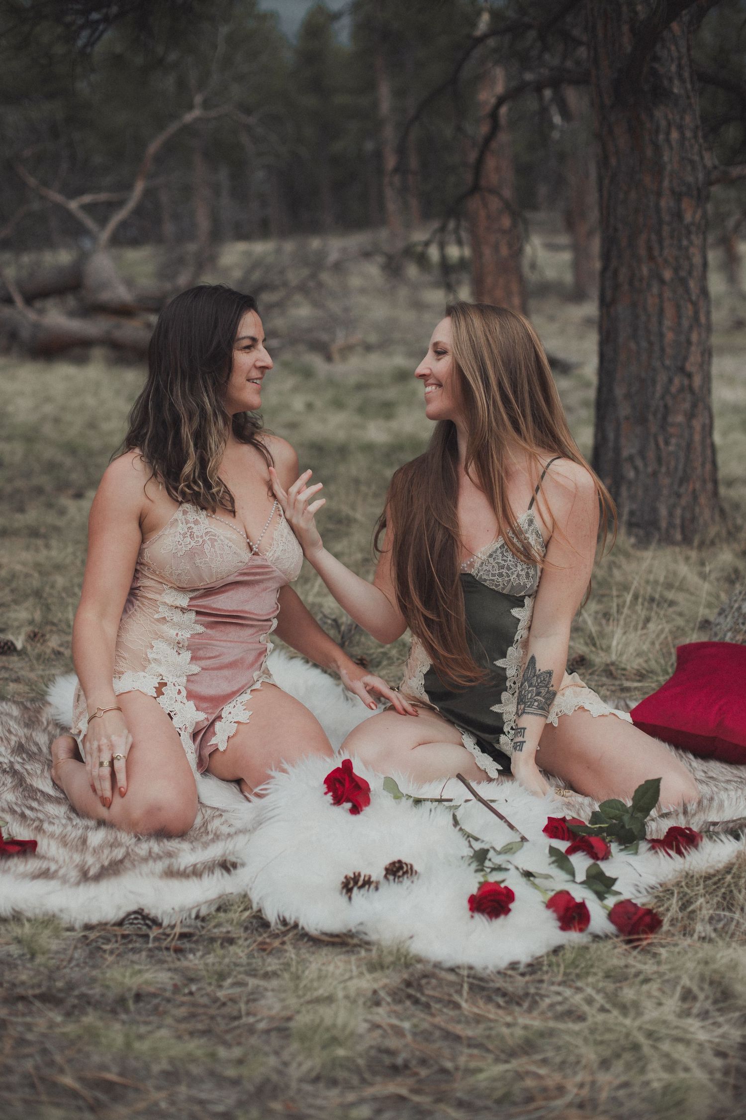 Two women in elegant dresses sitting on white blanket with red roses scattered around in forest setting.