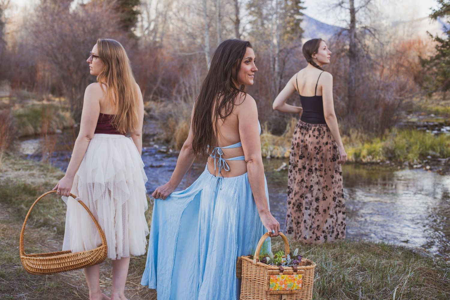 Three women in flowing dresses carrying baskets by riverside at sunset.