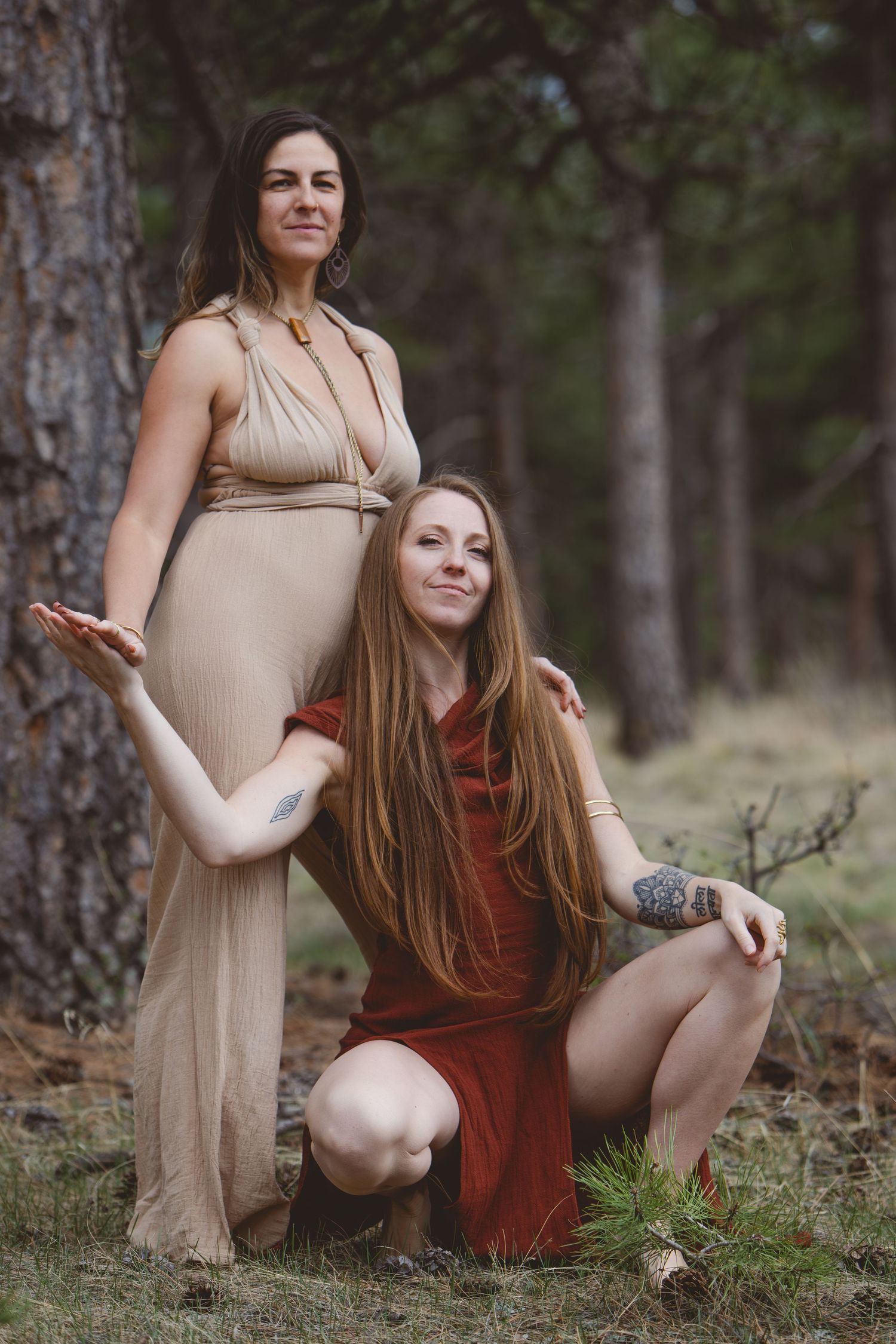 Two women in elegant dresses pose artistically against a pine tree in a forest setting.