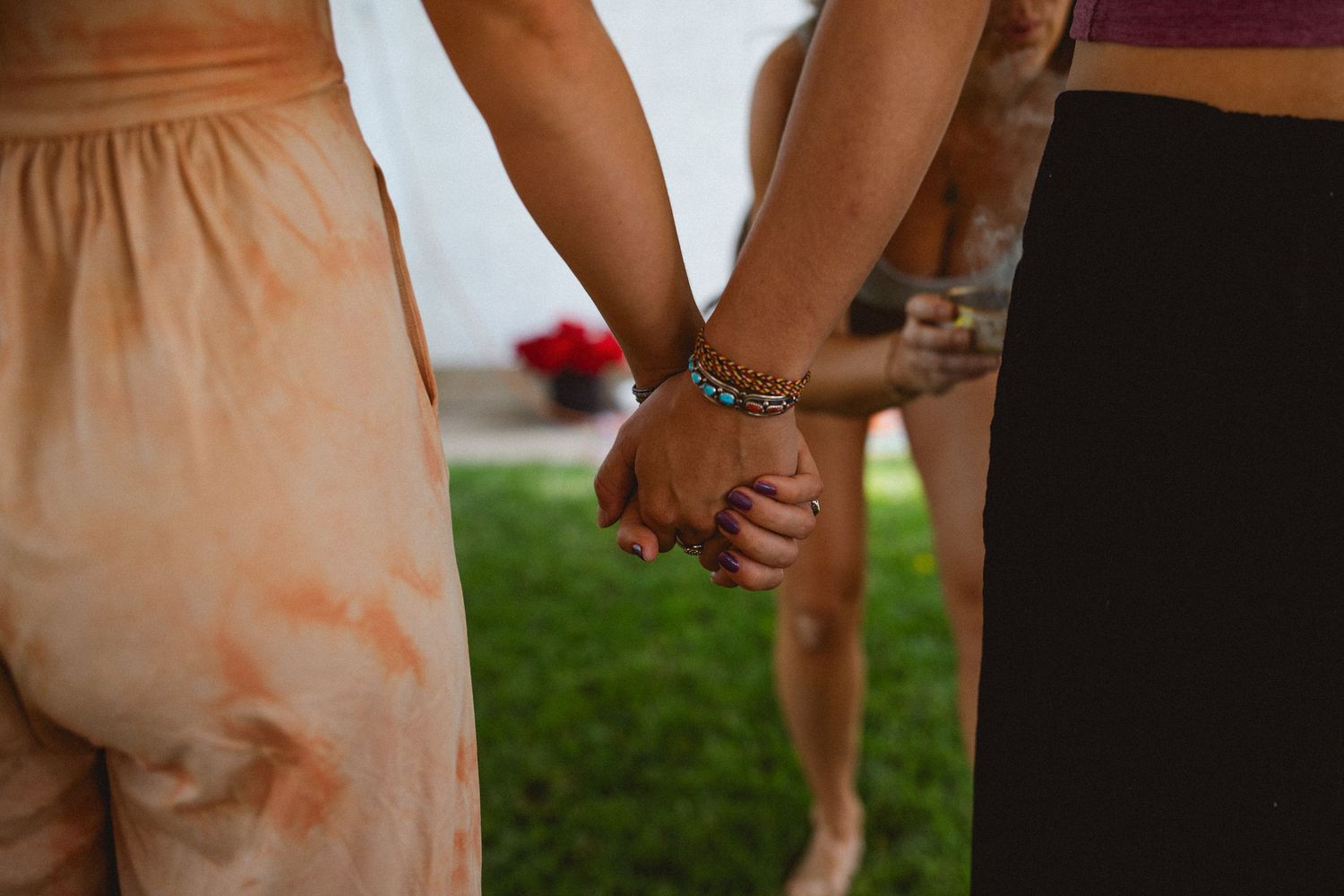 Close up of two people holding hands while standing on grass wearing summer outfits and bracelets.