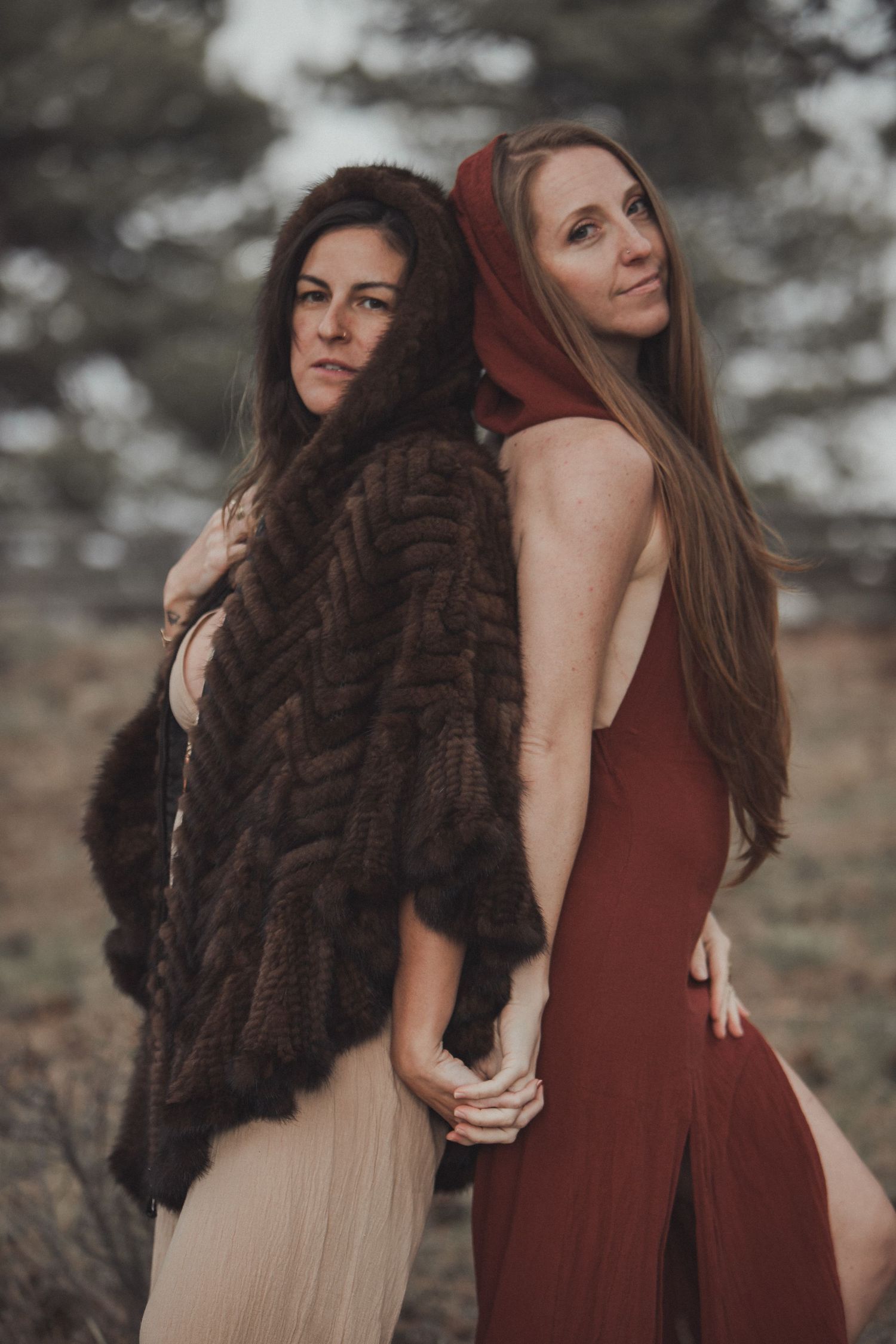 Two women in long dresses pose together outdoors, one wearing a brown fur coat while the other wears a burgundy dress.