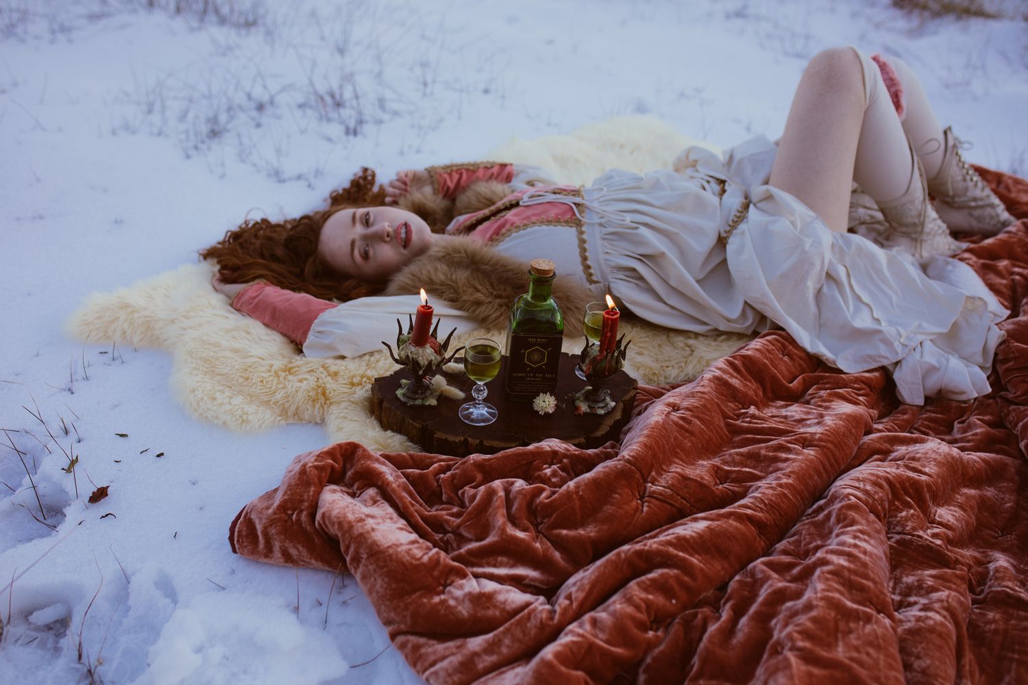 Cozy winter picnic setup with bottles and candles on red blanket in snow.