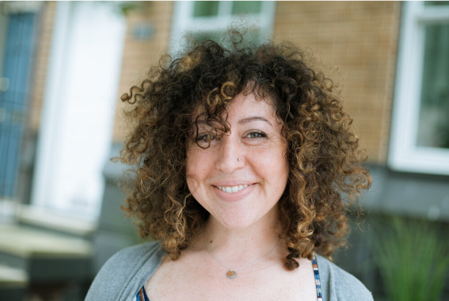 Person with curly hair smiles outdoors near brick building.