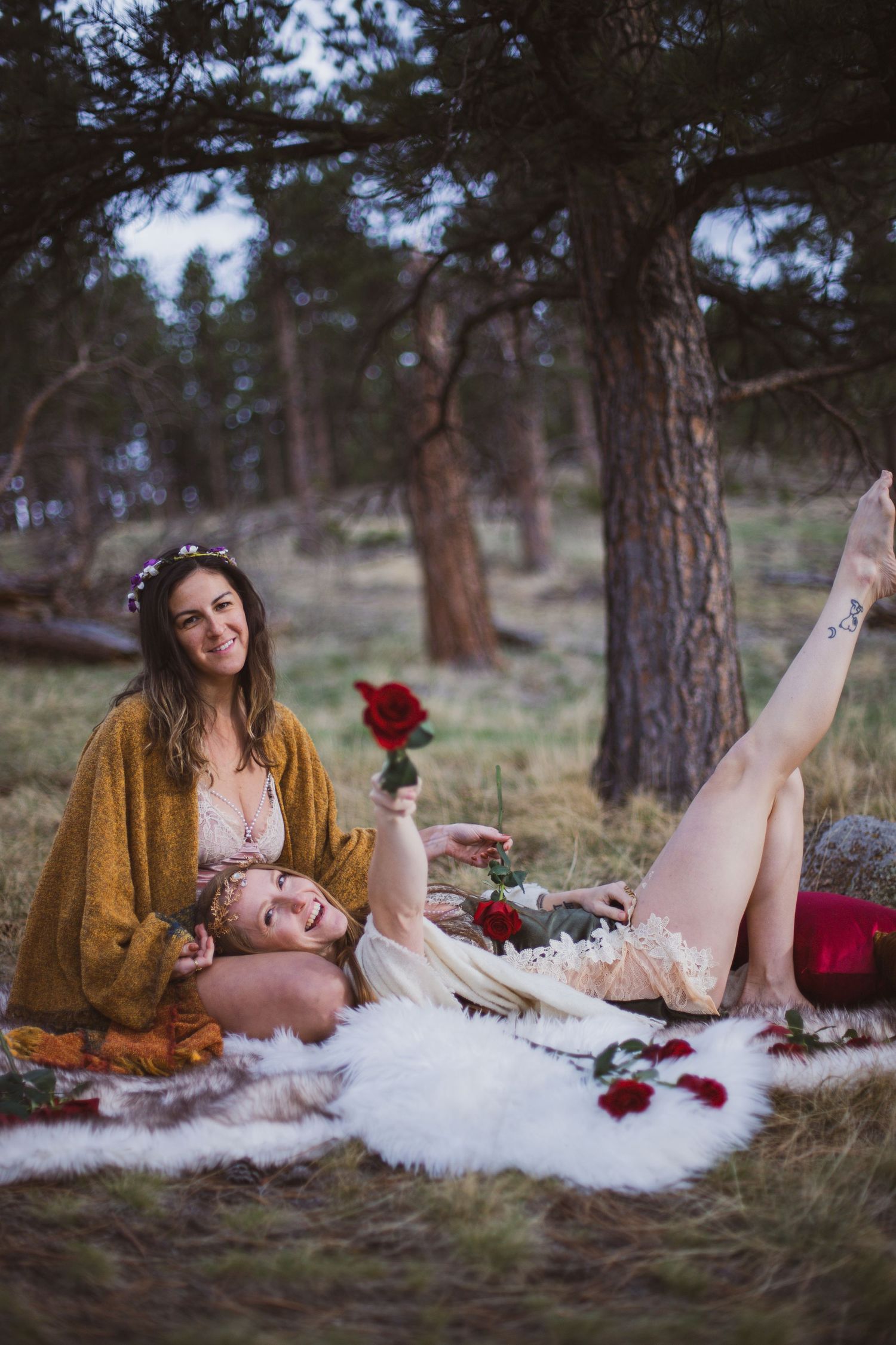Dreamy forest scene with two people lounging on white fur blanket surrounded by red roses and pine trees.
