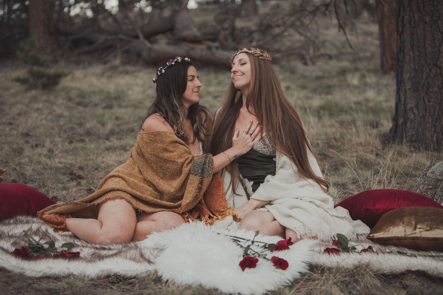 Two friends in bohemian dresses share laughter on white blankets with scattered roses in a moody forest setting.