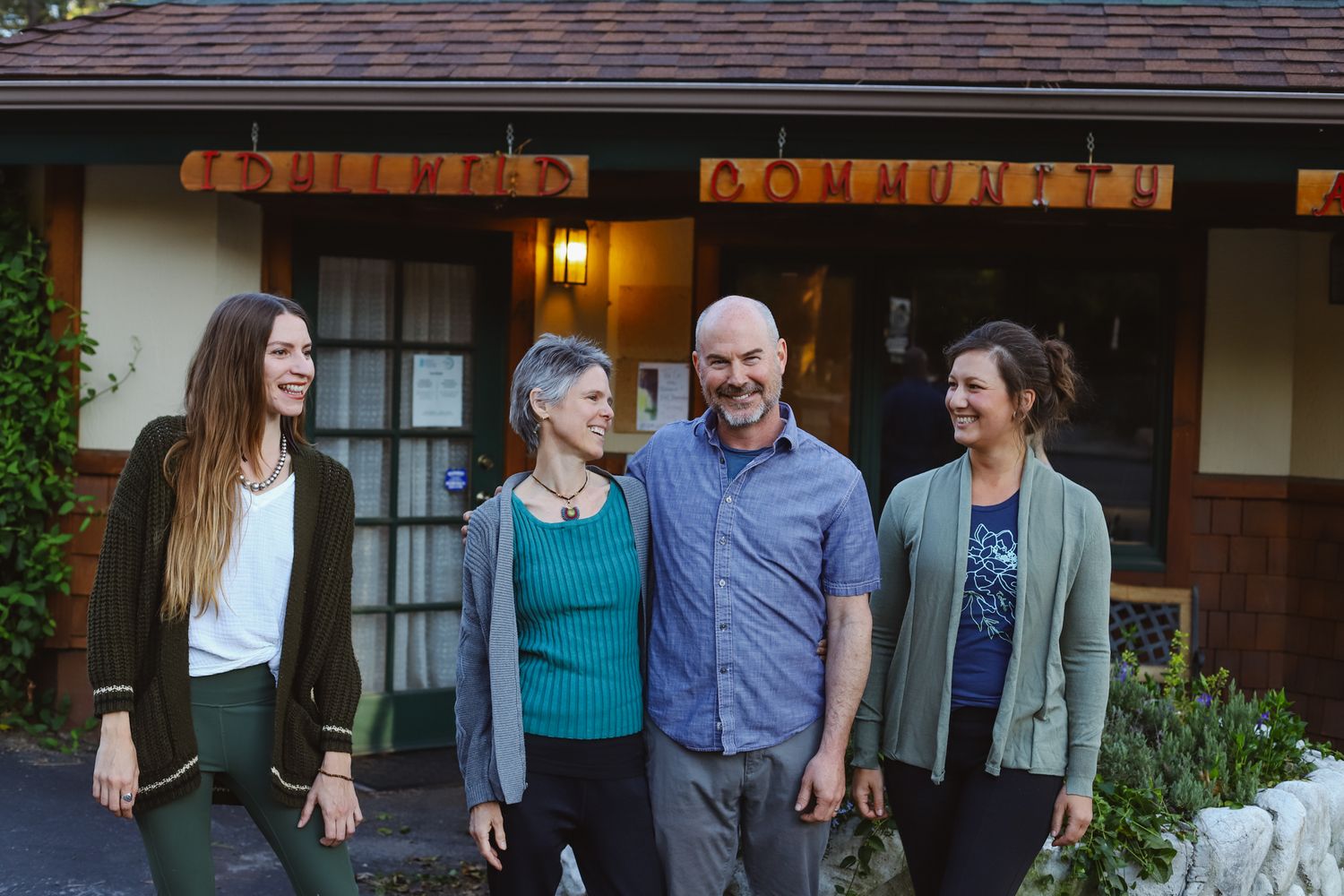 Four people stand together smiling in front of a house with warm lighting at dusk.