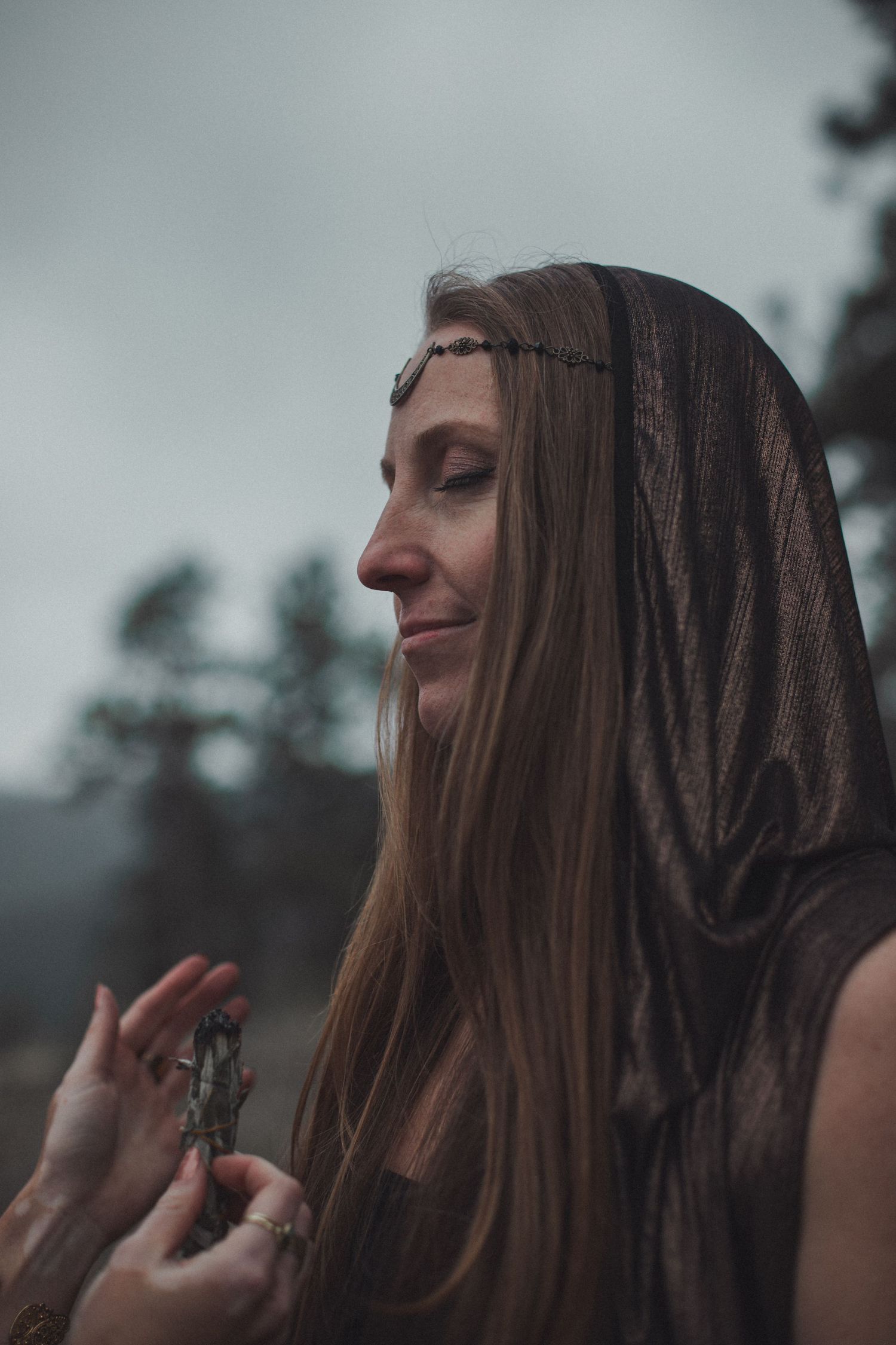 A person with long brown hair wearing a dark hooded top stands against a moody gray sky in a contemplative pose.