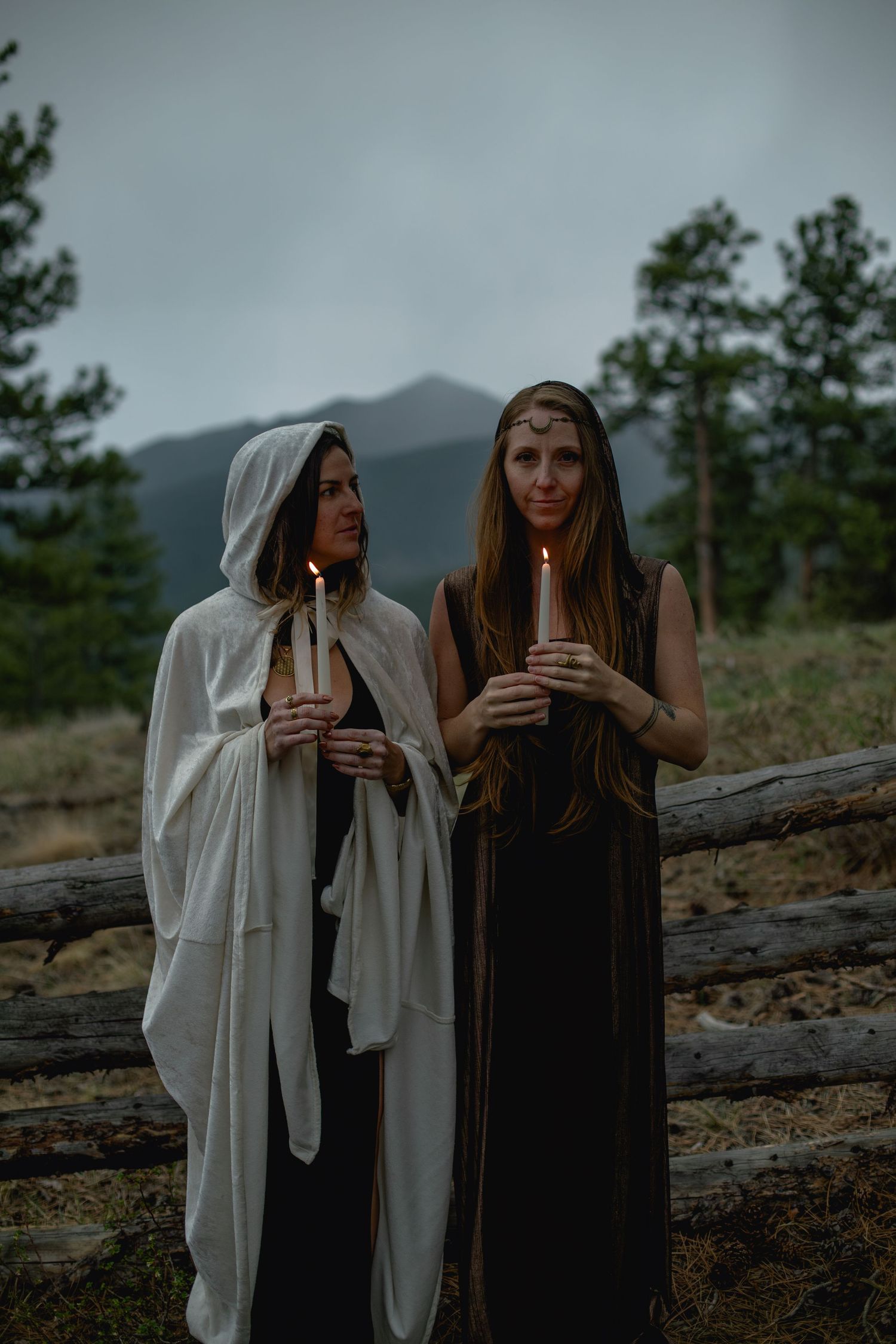 Two people in medieval style cloaks stand on a mountainside at dusk holding candles against a moody cloudy sky.