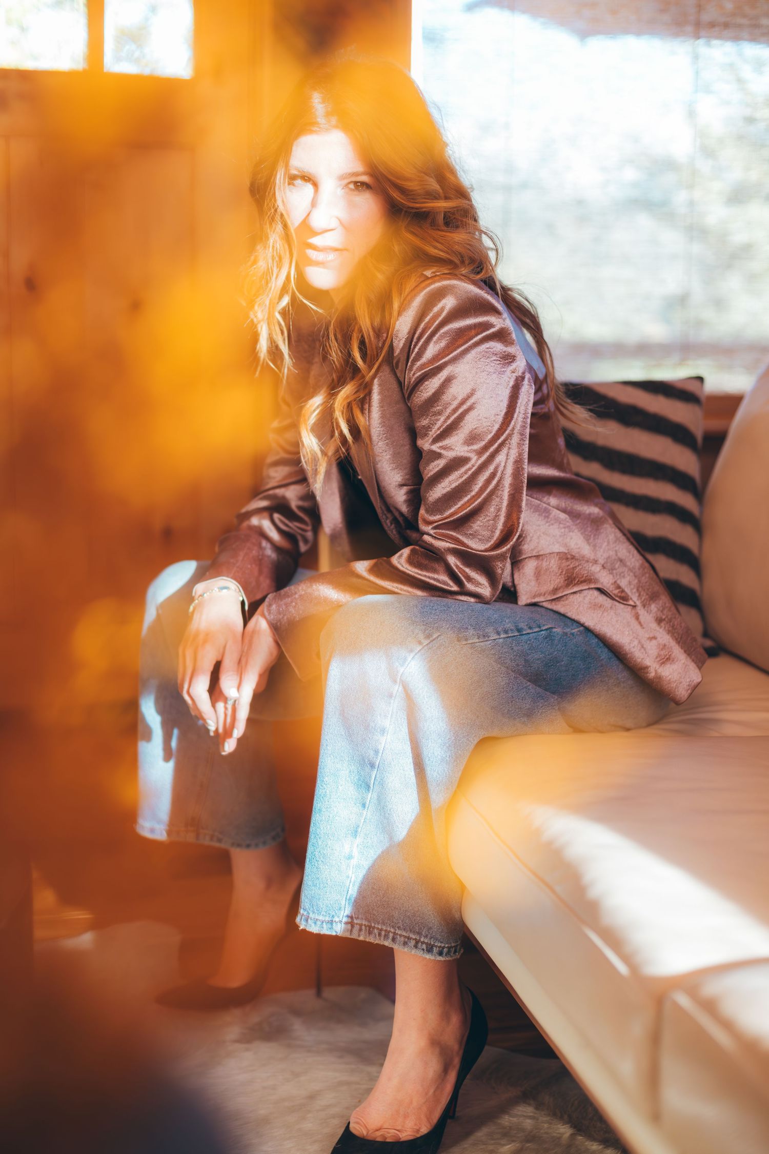 Woman in brown leather jacket and jeans sits on couch with warm lighting and soft focus effects.