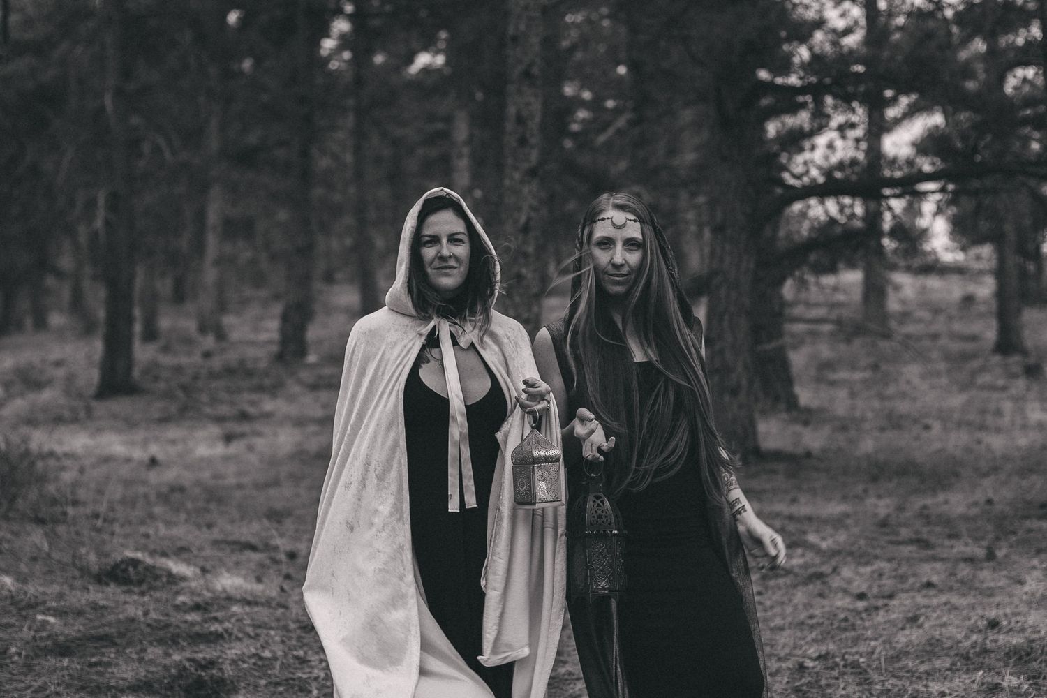 Two figures wearing dark and light cloaks walk together in a moody forest setting captured in black and white.