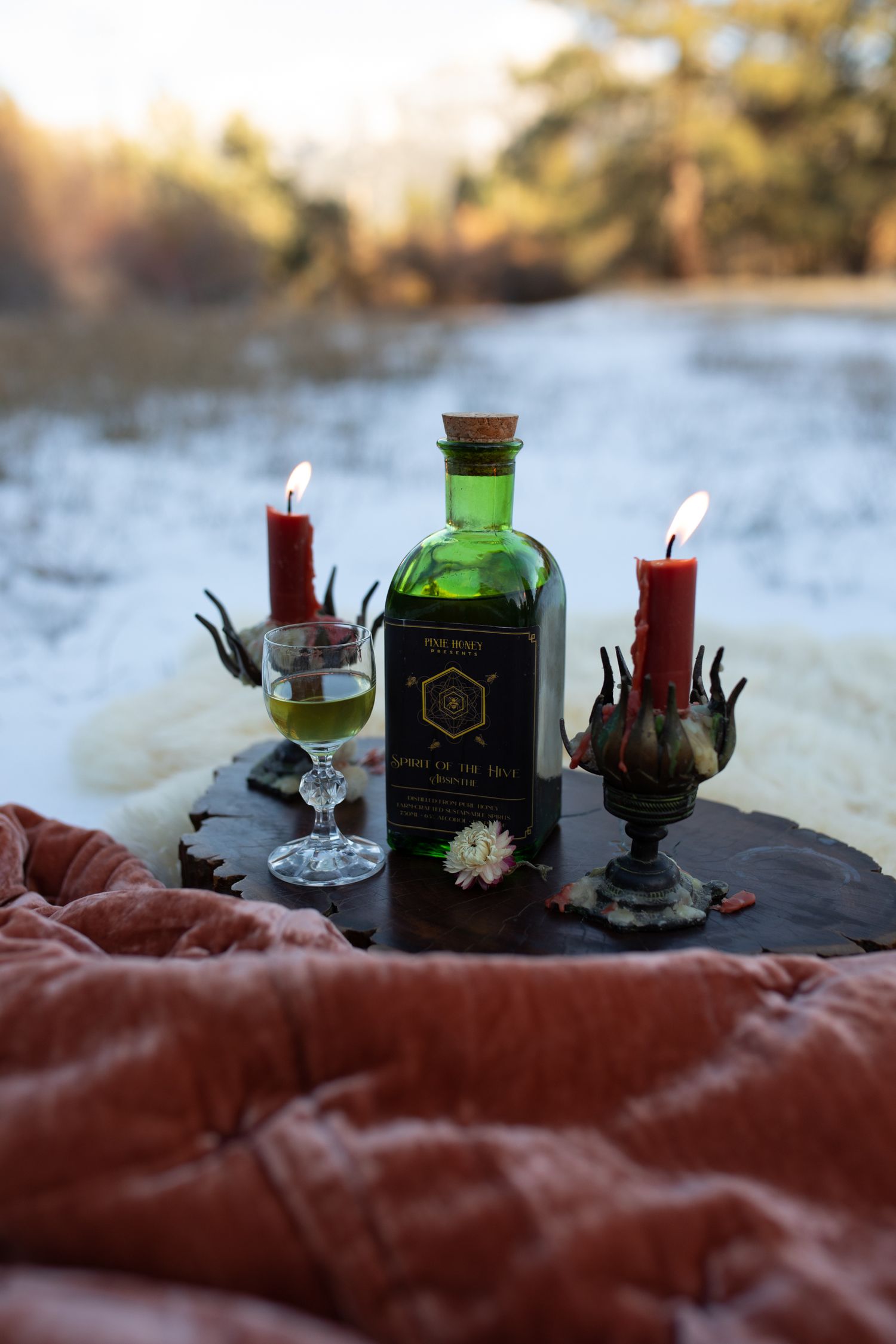 Green bottle and red candles arranged on blanket in snowy outdoor scene.