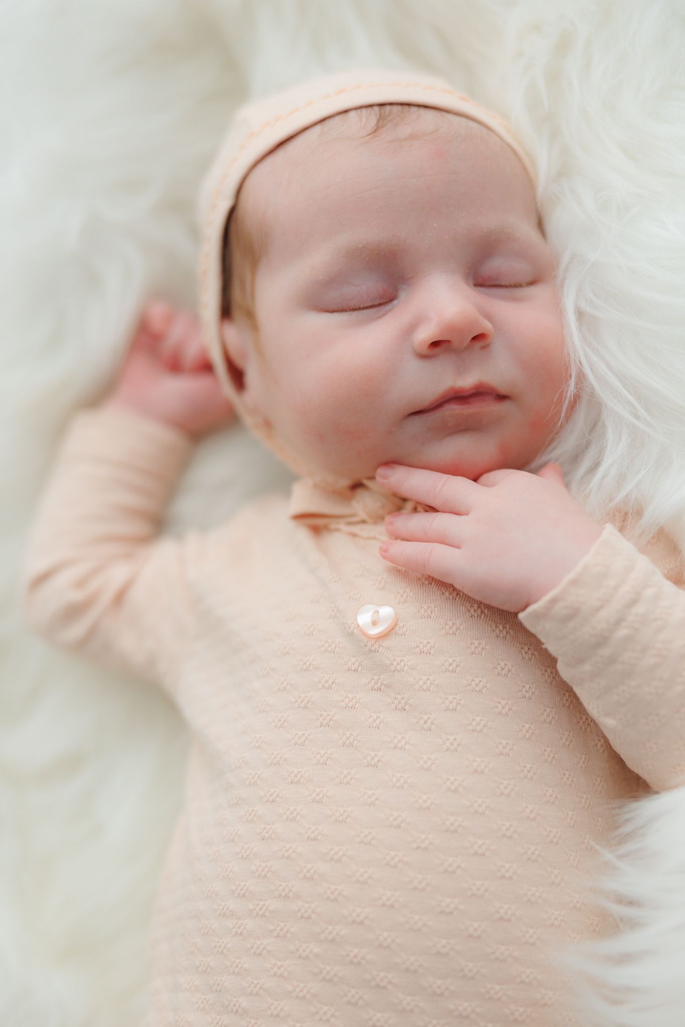 Sleeping newborn baby in pink outfit and headband resting peacefully on white fluffy blanket.