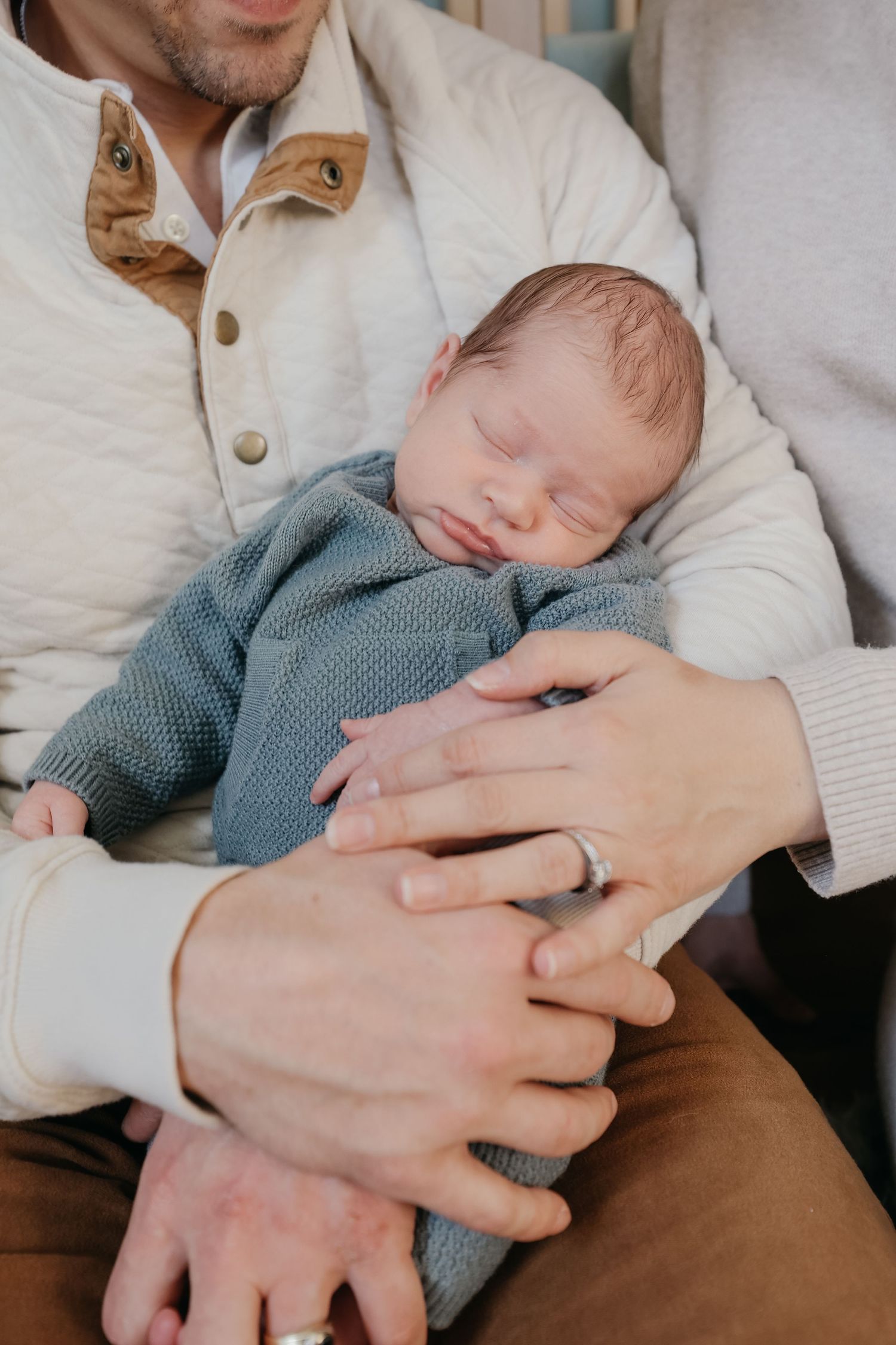 Close up of sleeping newborn being held in parents' hands wearing blue outfit.
