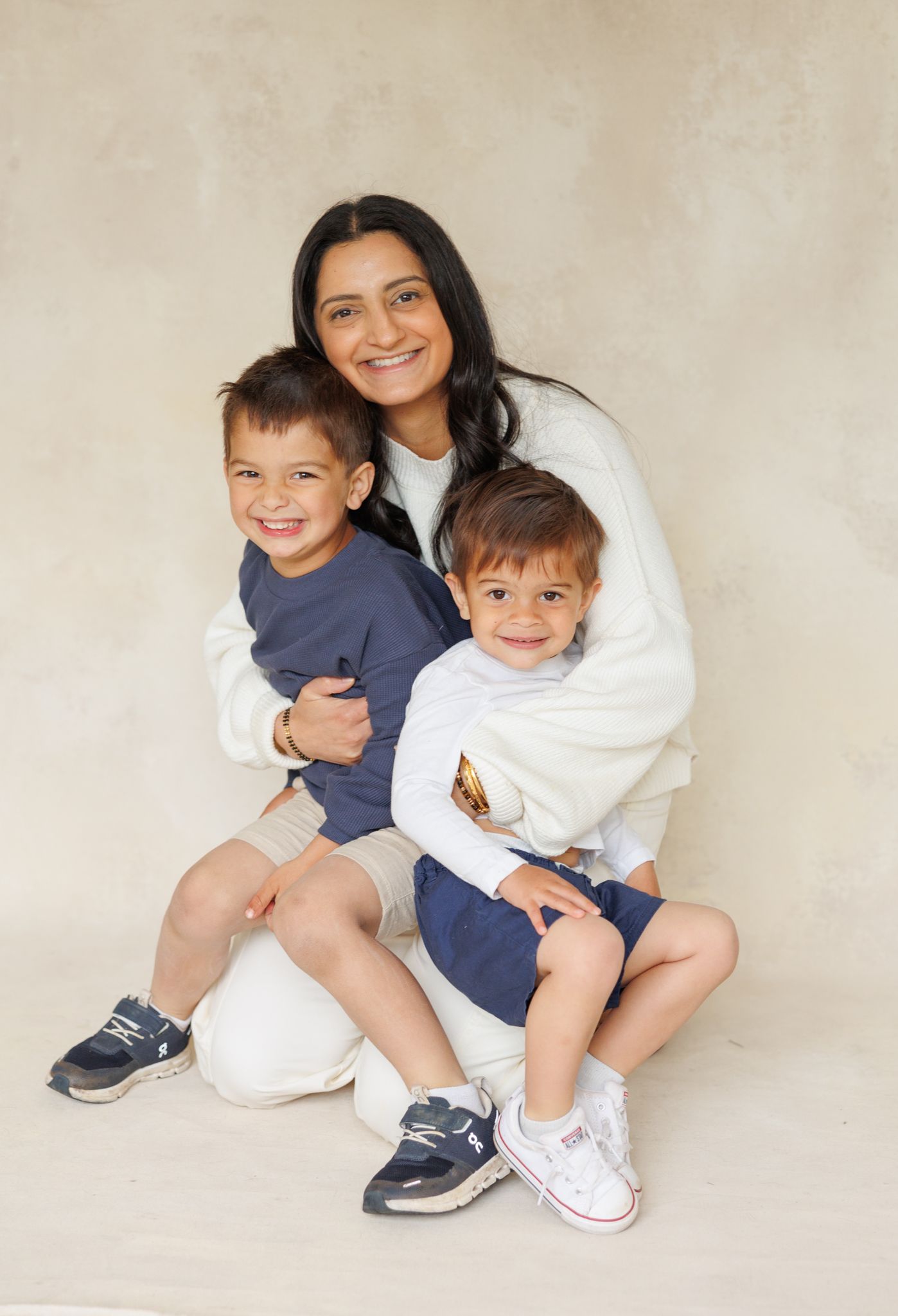 A smiling woman hugs two young boys in navy shorts, posing together for a portrait against a beige background.