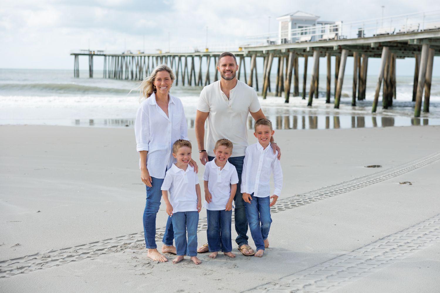 Family in white and blue outfits walking along sandy beach near pier during sunset photo session.