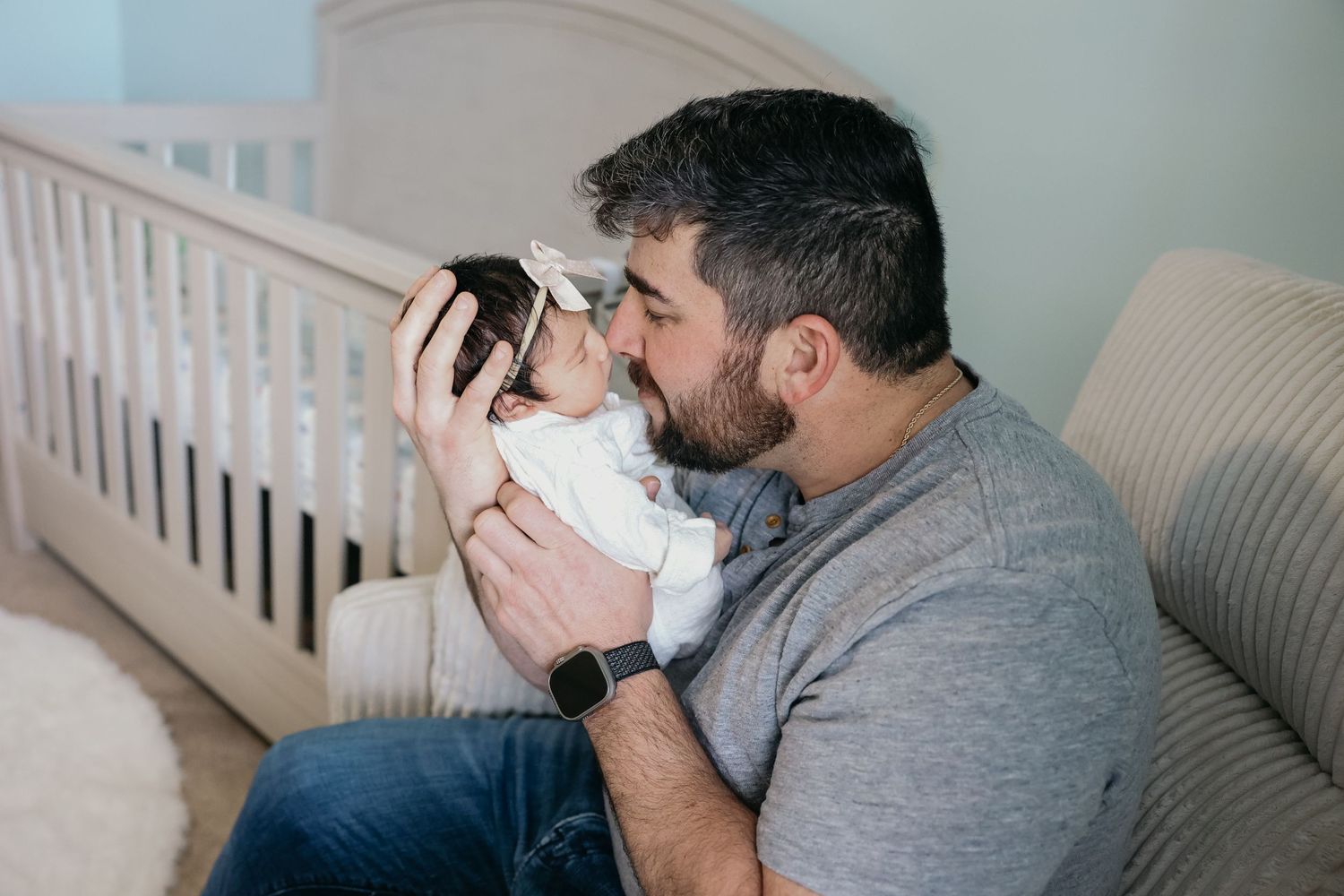 A person in a grey sweater tenderly holds and kisses a newborn baby in a white onesie next to a crib.