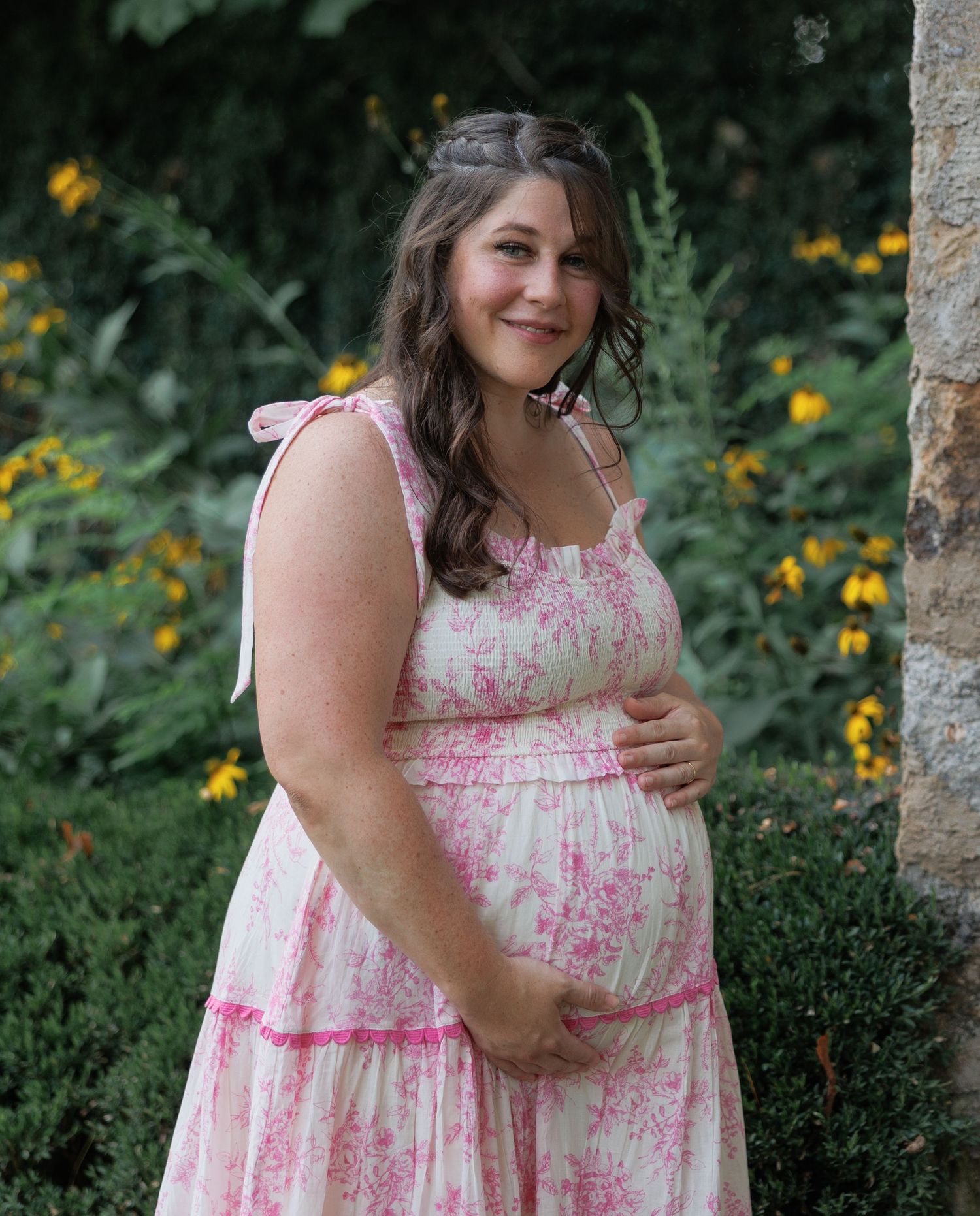 Someone in a pink and white floral sundress poses outdoors against a garden backdrop with yellow flowers.