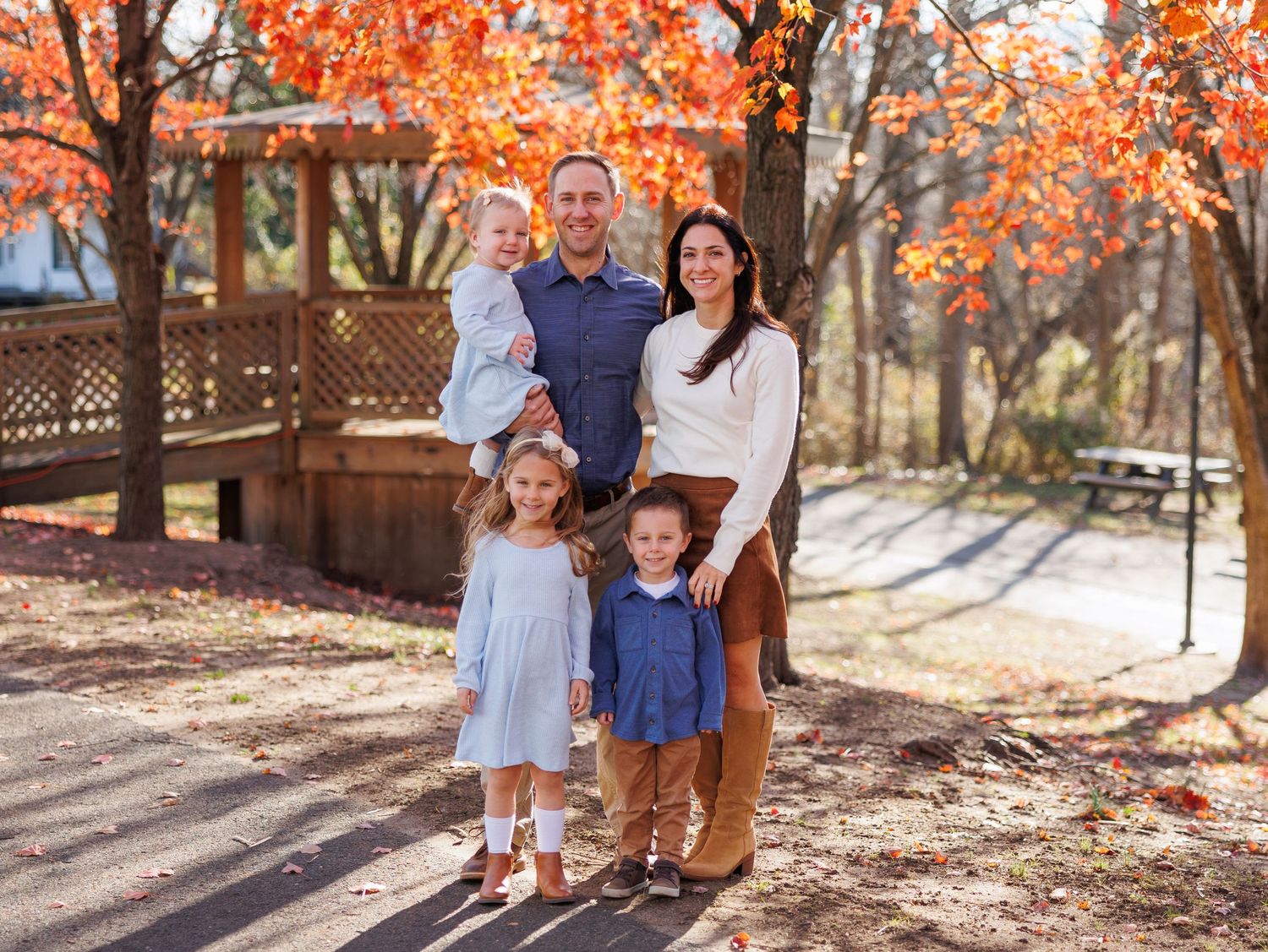 A family stands together outdoors during autumn with vibrant orange fall foliage and a wooden gazebo in the background.