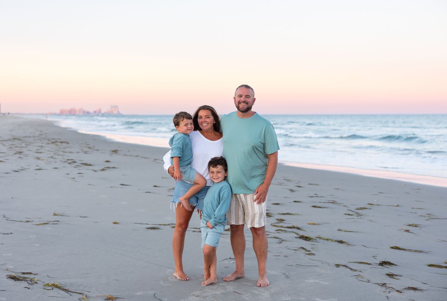 Family of four posing on a sandy beach at sunset with calm ocean waves and a pastel sky in the background.