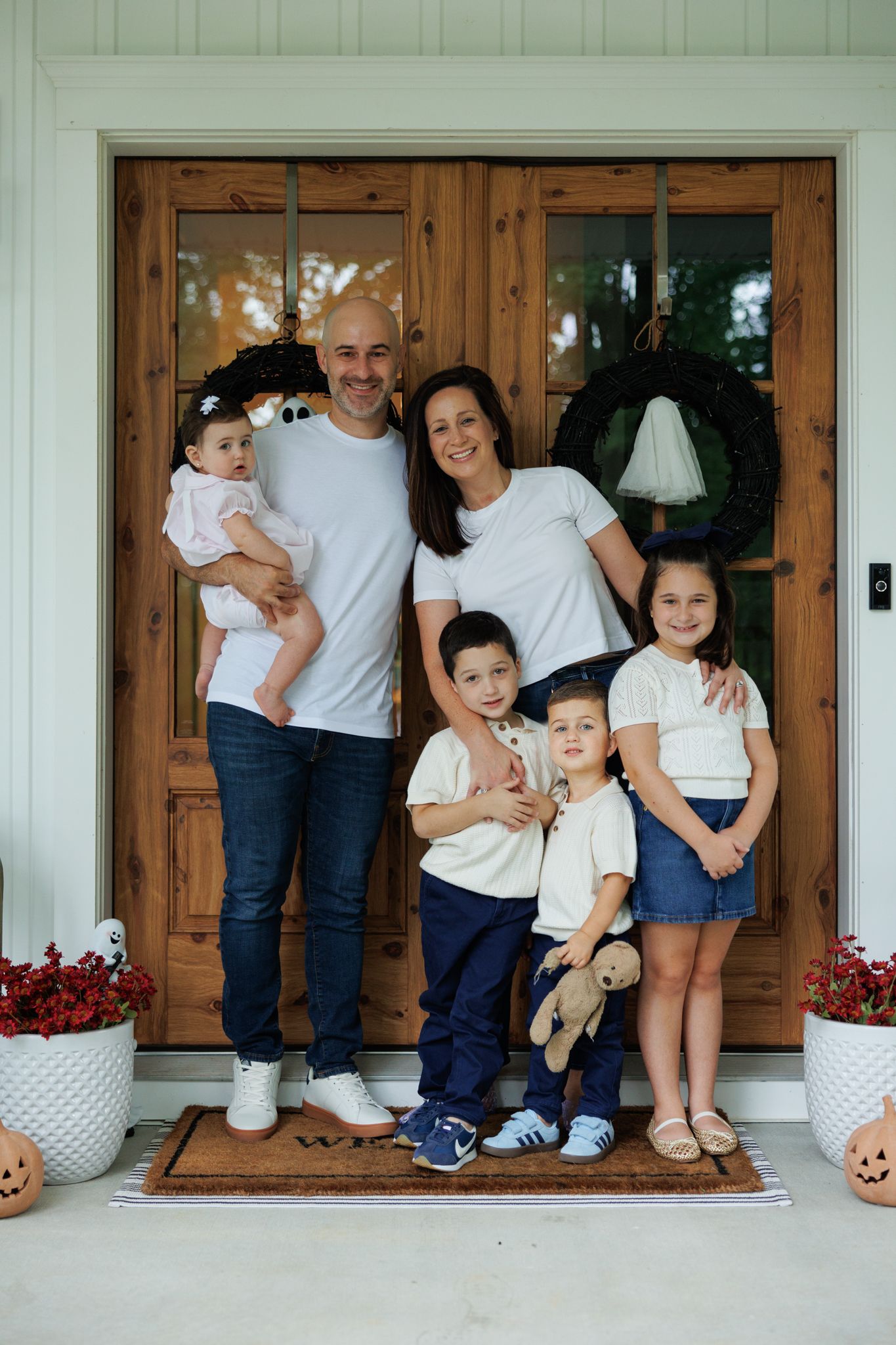 Happy family of five posing on front porch with Halloween decorations, wearing white tops and jeans.
