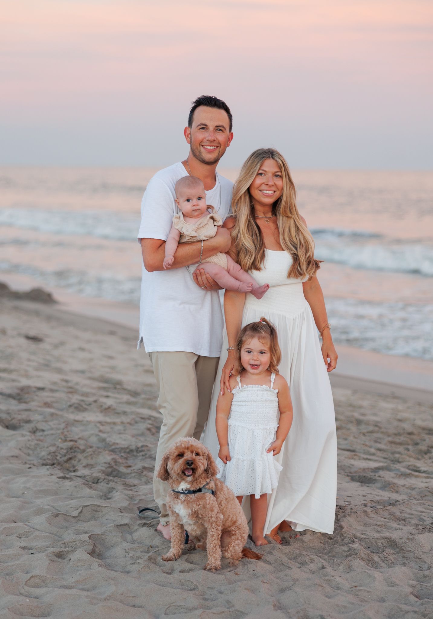 Family of four with a dog posing on a sandy beach at sunset, wearing coordinating white and neutral outfits.