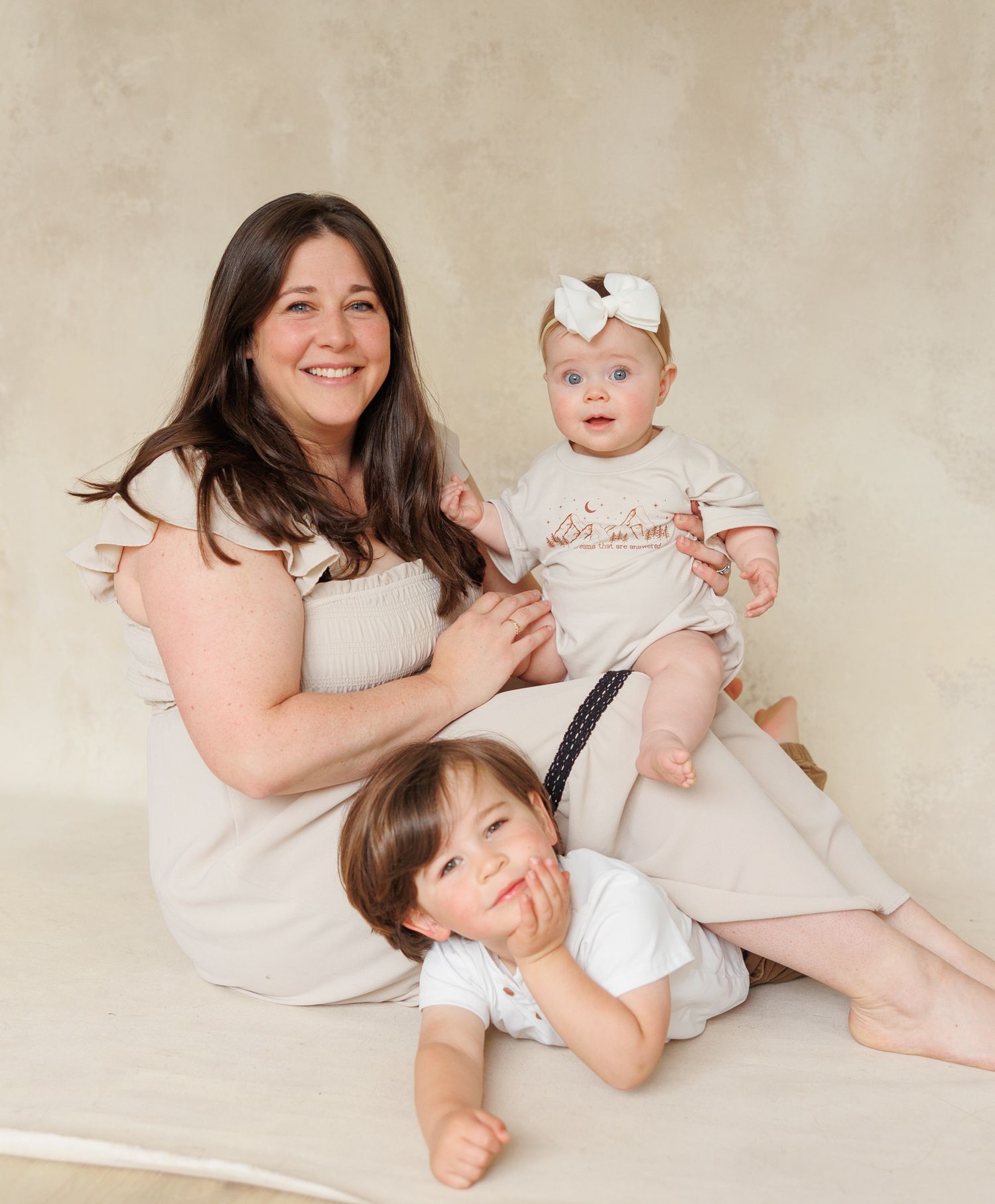 A woman sits smiling with two young children, a toddler and a baby girl in a bow, in a soft studio portrait.