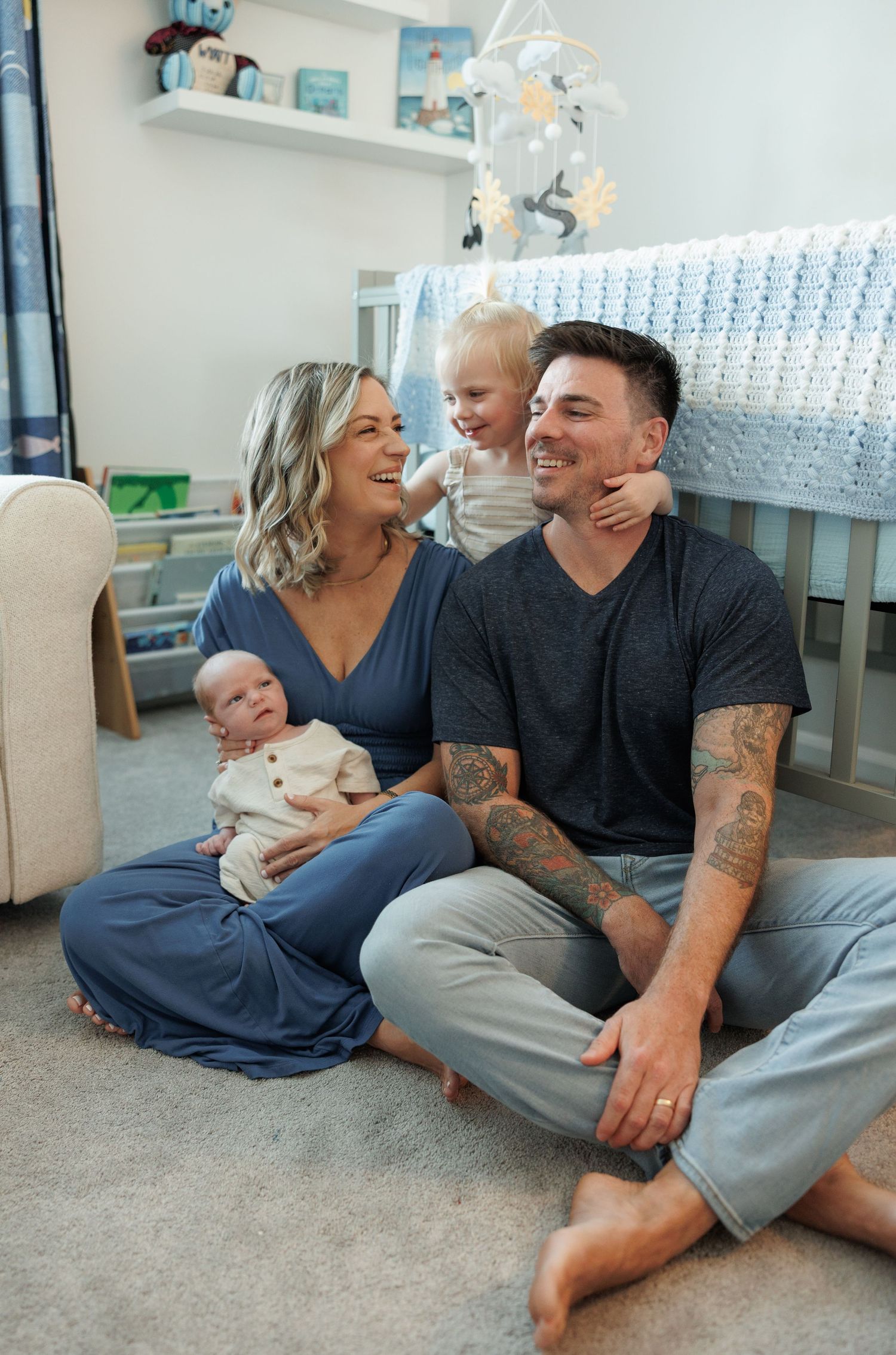 A happy family sits on a nursery floor, mom holding a newborn, toddler perched on tattooed dad's shoulders.