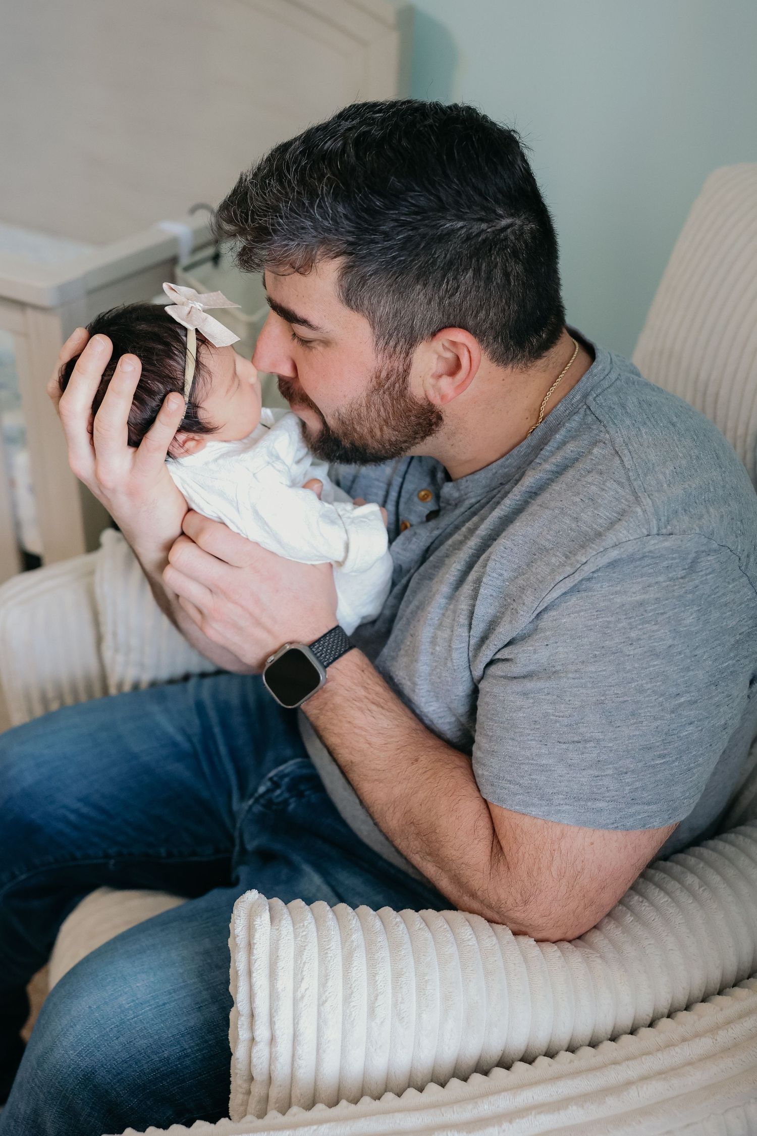 A tender moment captured as a person in gray holds and kisses a newborn baby in white clothing while sitting in a chair.