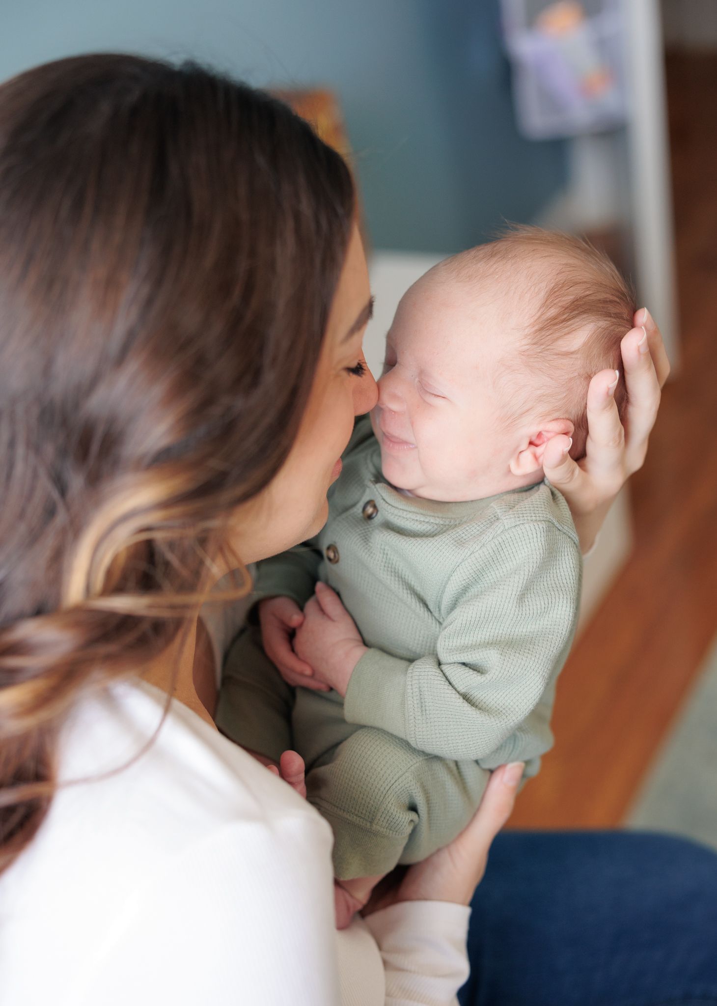 A mother tenderly kisses her newborn baby dressed in a sage green romper while cradling them in her arms.