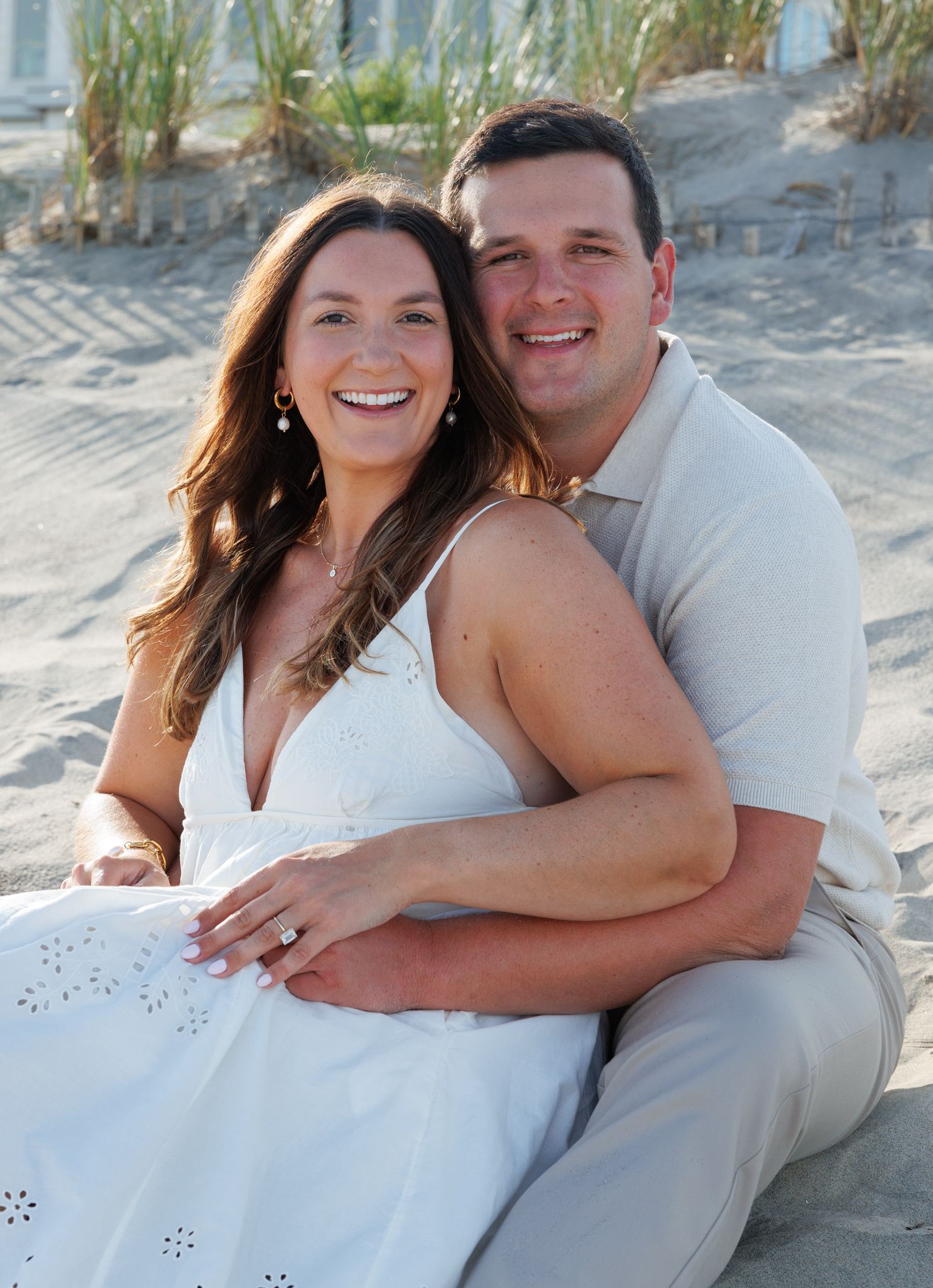 Happy couple sitting on beach sand, woman in white dress showing engagement ring, man in beige polo shirt, smiling outdoors.