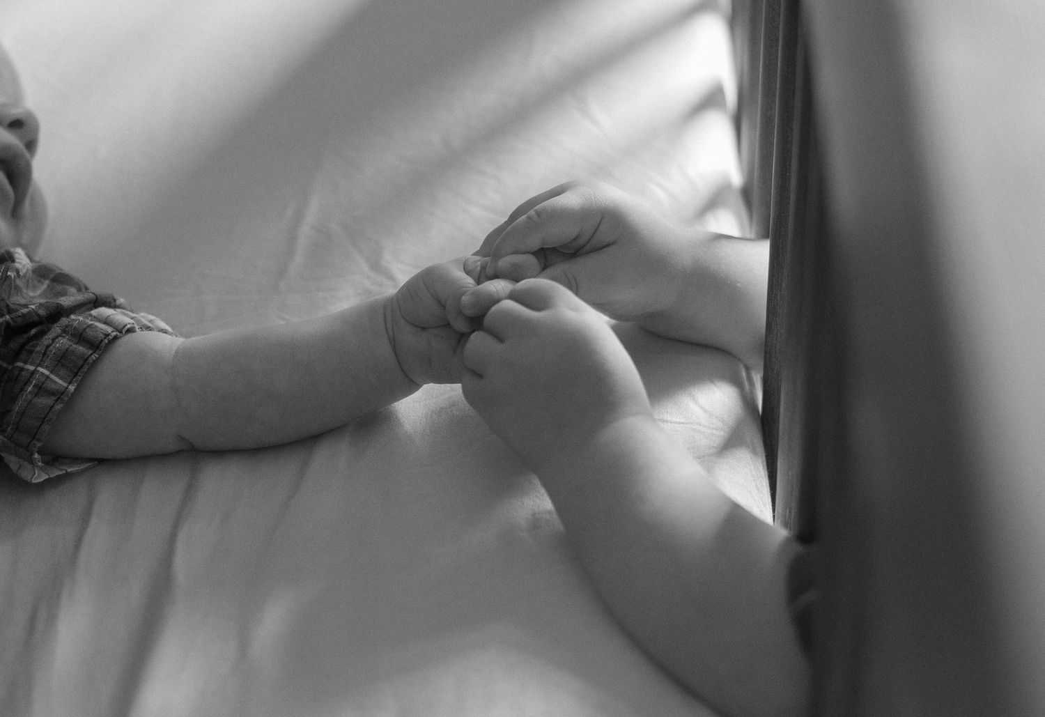 Black and white photo of a baby and adult holding hands on a white bed sheet.