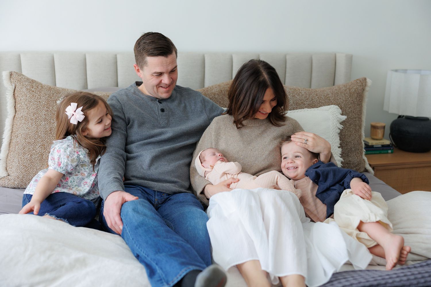 Happy family of five sitting on a bed, parents with three young children including a newborn, in a cozy bright bedroom.