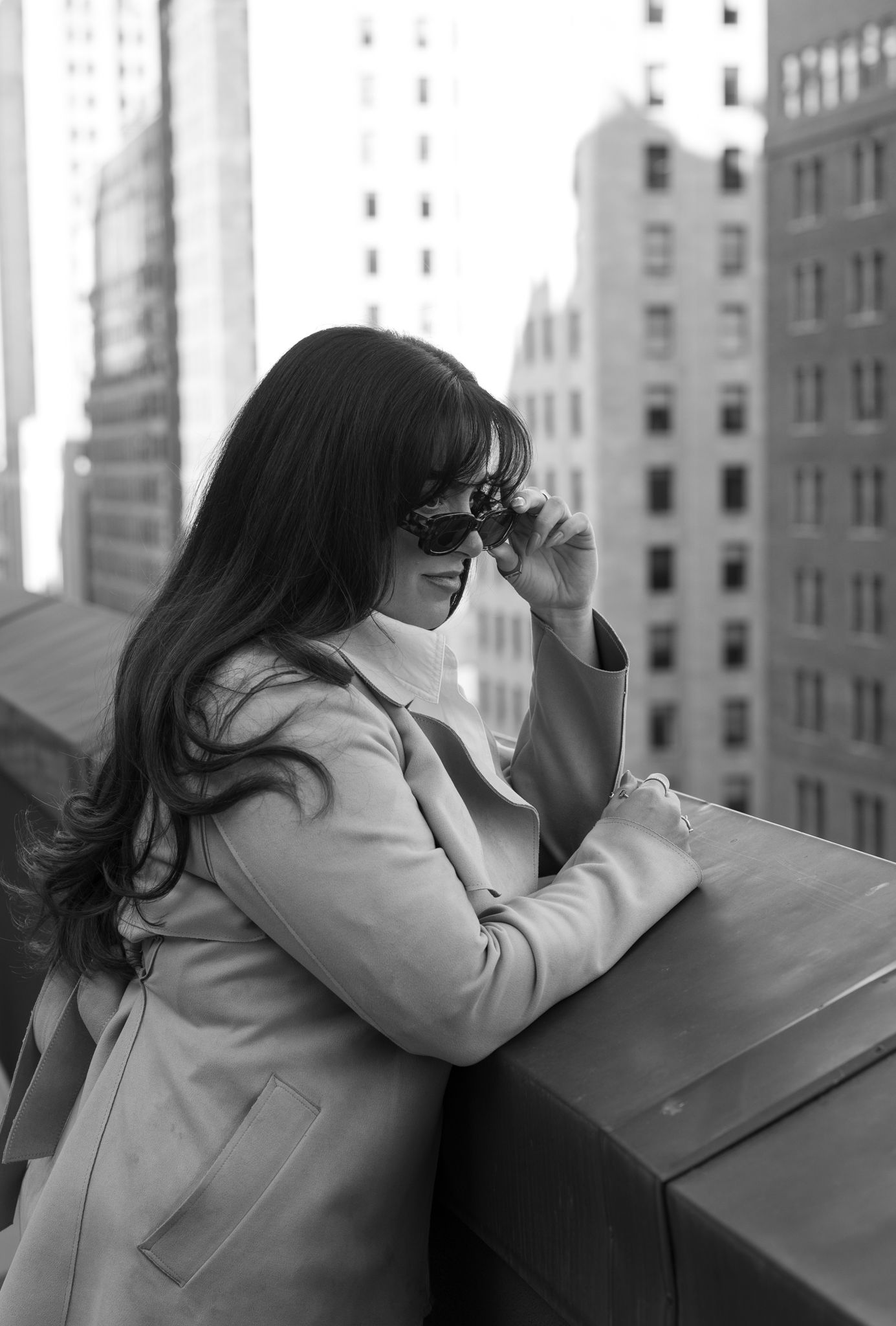 Black and white photo of a woman in a coat adjusting sunglasses on a city rooftop.