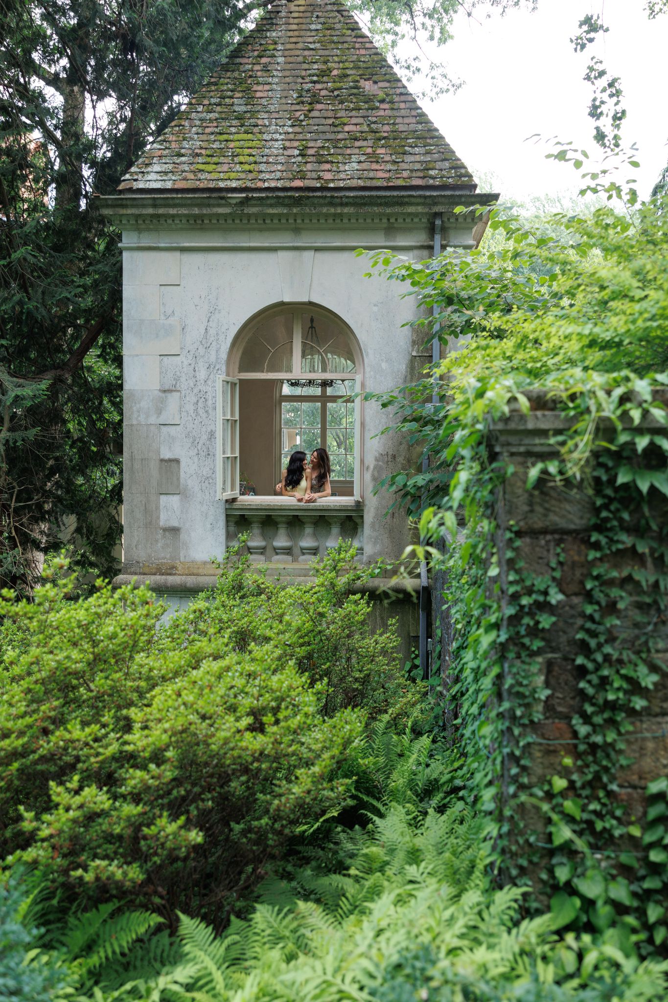 Two people sit in an arched window of a moss-covered stone garden pavilion surrounded by lush green foliage.