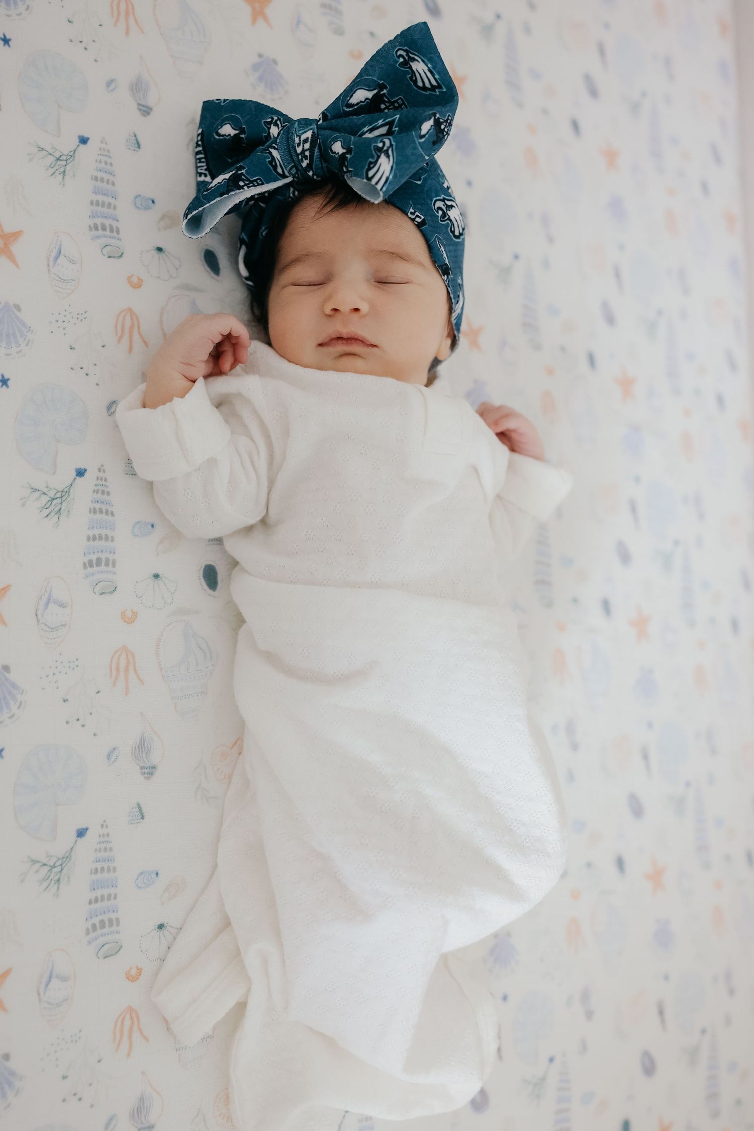 Sleeping newborn in white outfit wearing large blue bow headband on floral print background.